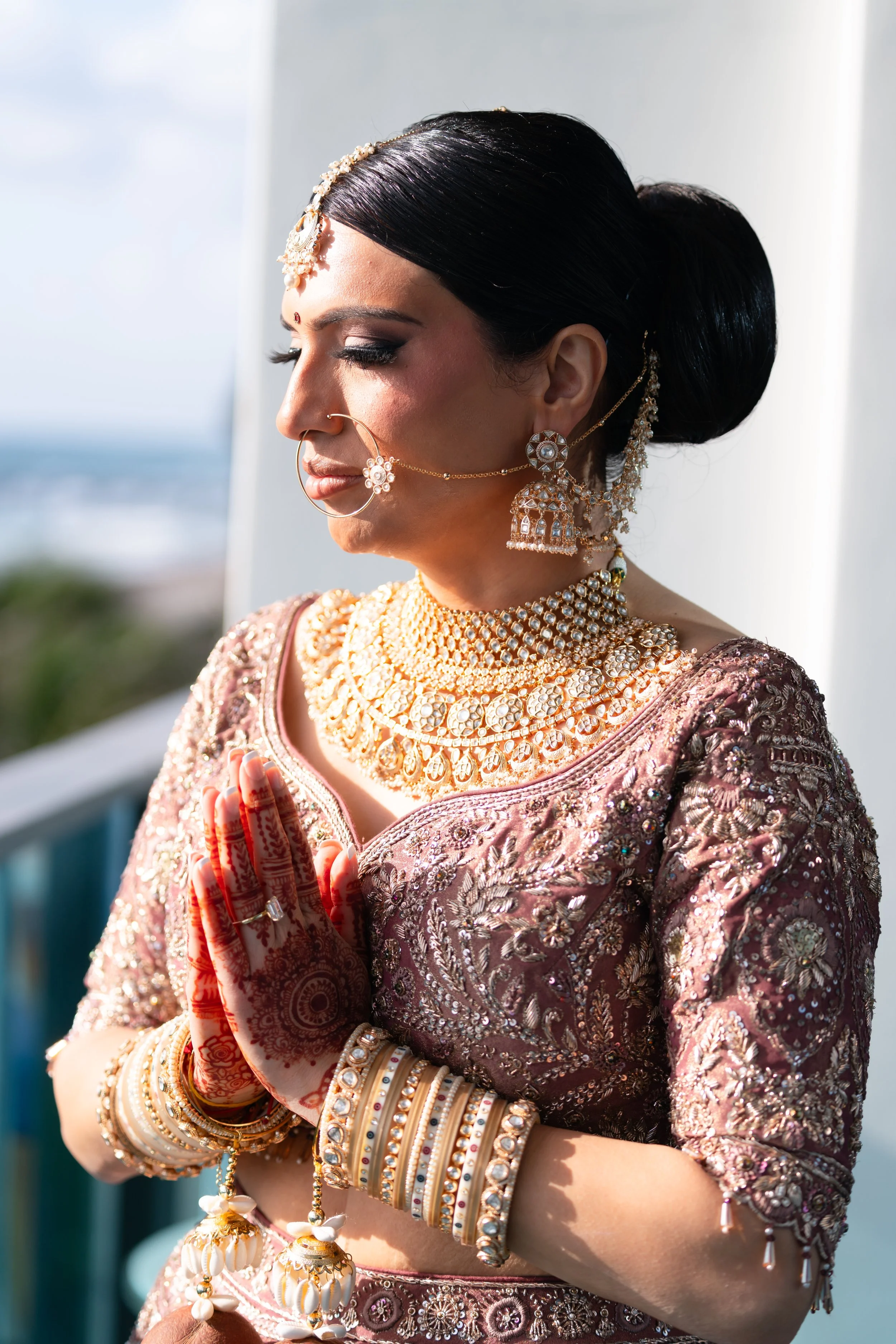 A woman dressed in traditional Indian attire, adorned with jewelry and henna, with her hands pressed together in a prayer pose, standing on a balcony with a view of the sky and distant landscape. South Florida Wedding Photographer, CA Wedding 