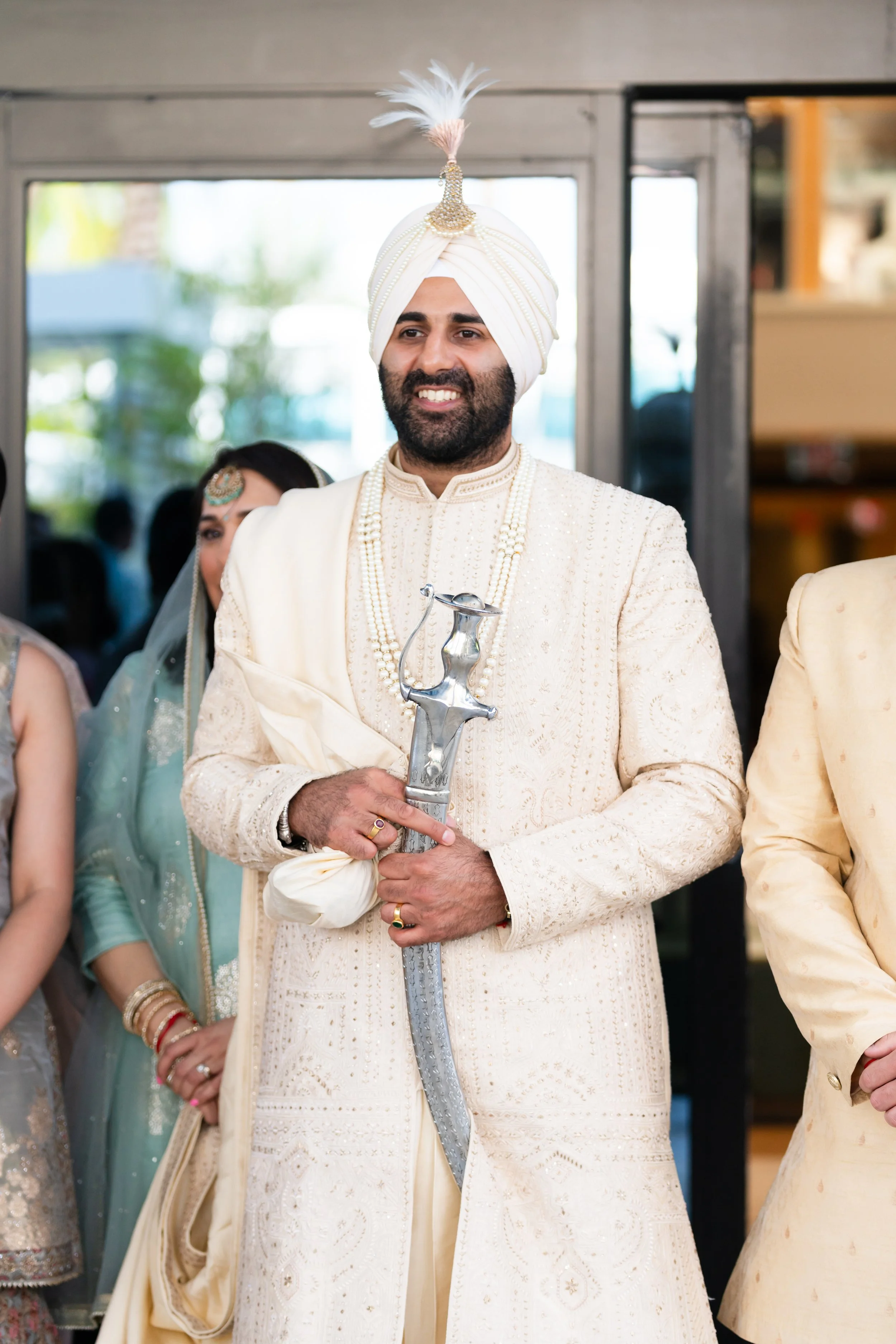 A groom wearing an ivory traditional Indian sherwani and turban, holding a sword, smiling during a wedding celebration. Indian Wedding, Cultural Wedding, Miami Wedding