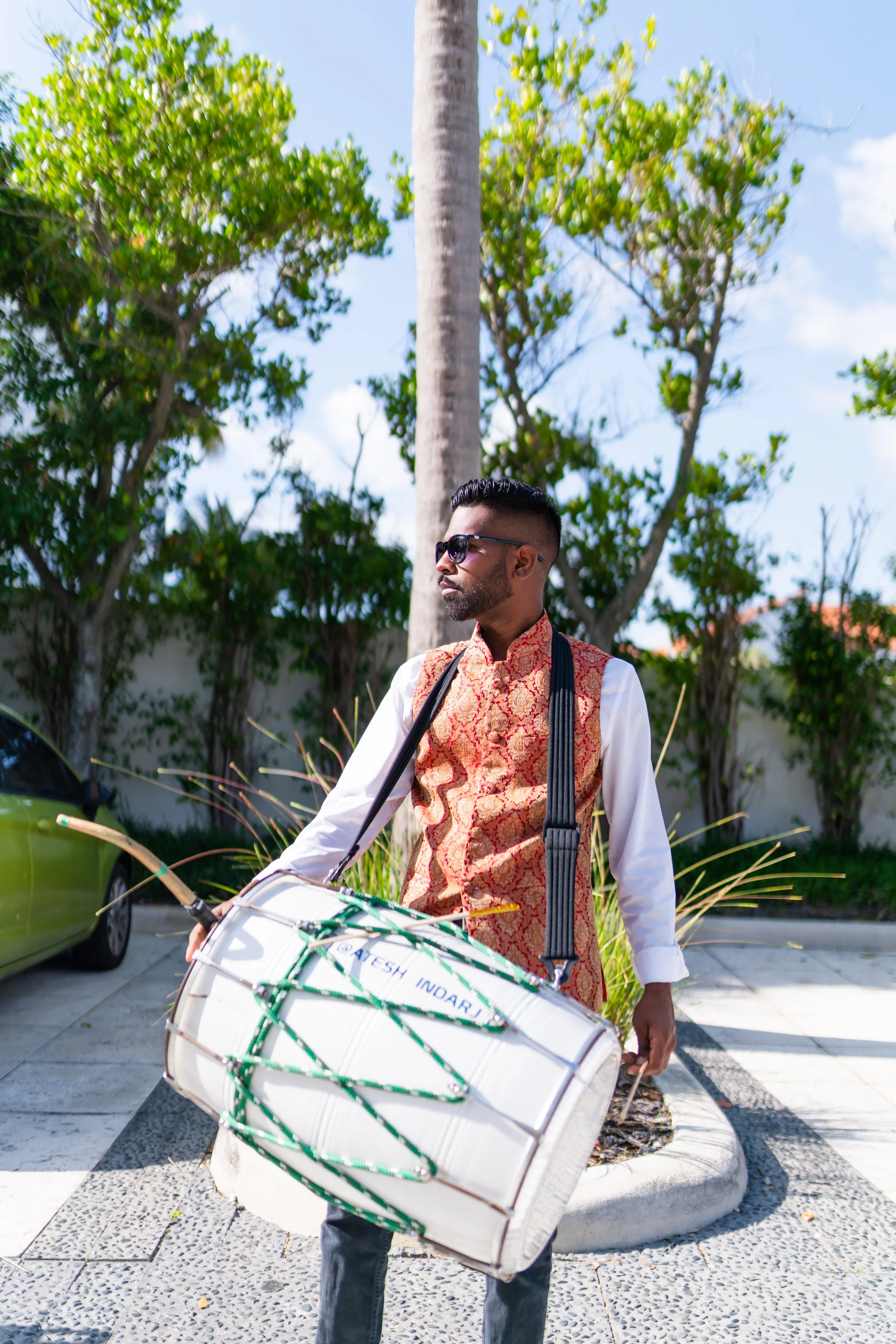 A man wearing sunglasses, a traditional red and gold vest, and a white shirt holding a large white drum with green ropes, standing outdoors near some trees and a parked car.