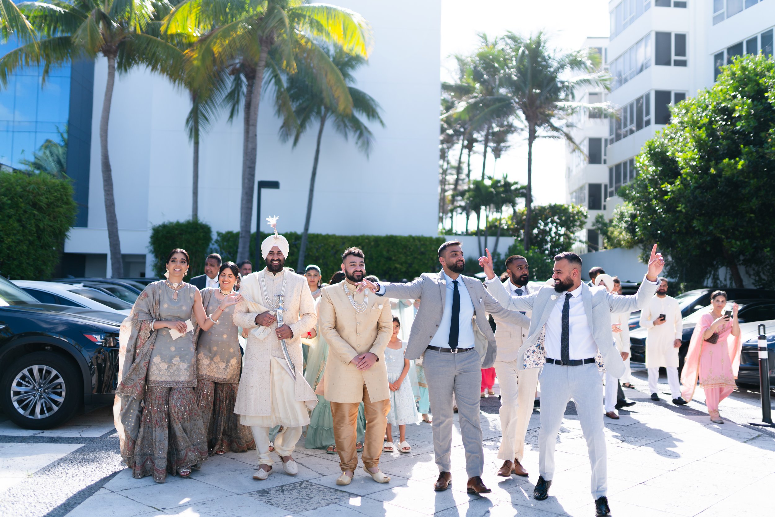 A group of people, dressed in traditional and modern clothing, celebrating outdoors in a sunny urban area with palm trees and tall buildings in the background.