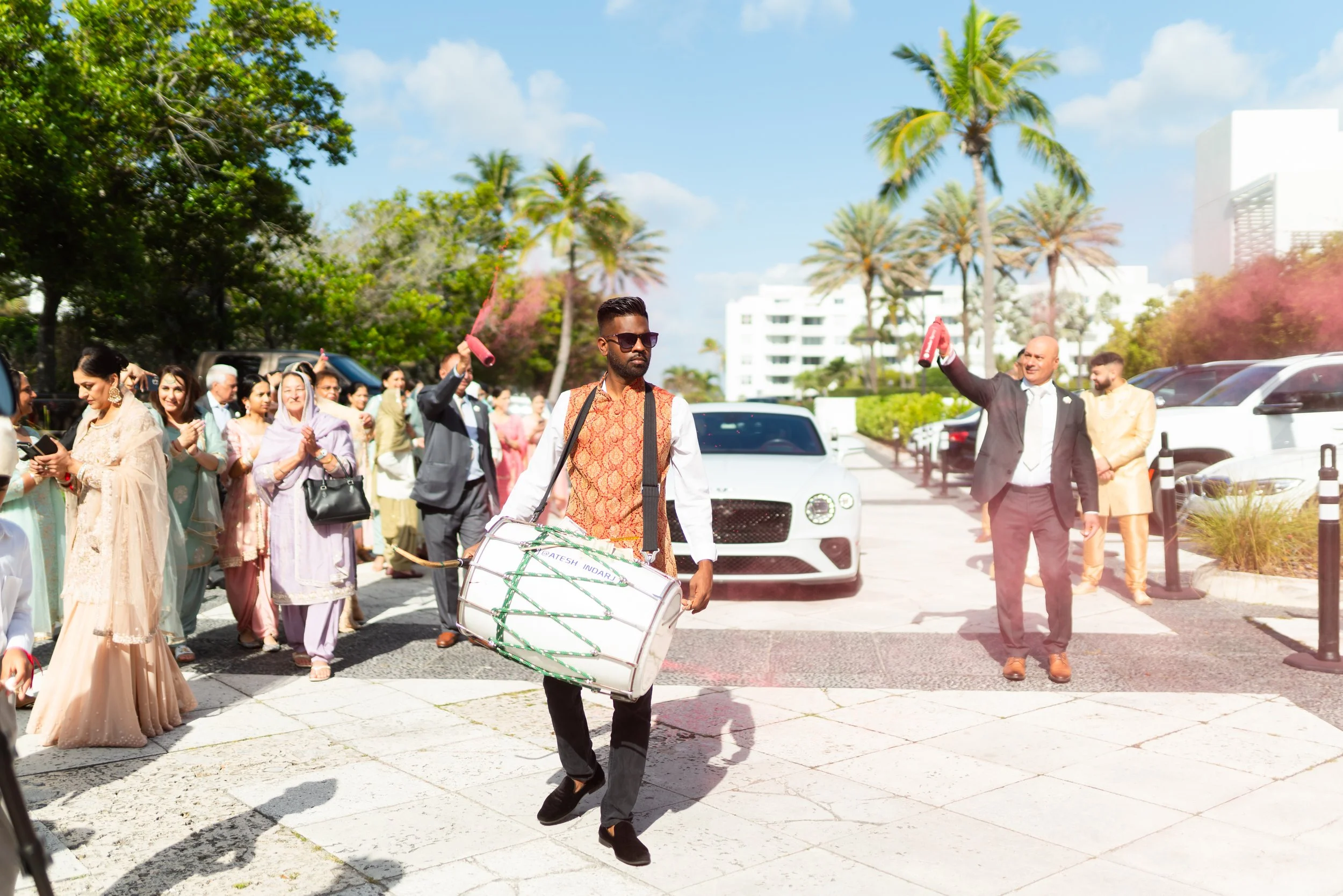 A man in traditional Indian attire carrying a large drum walks in front of a crowd of people, with some holding smoke devices, in an outdoor setting with a parking lot, trees, and a white building in the background.