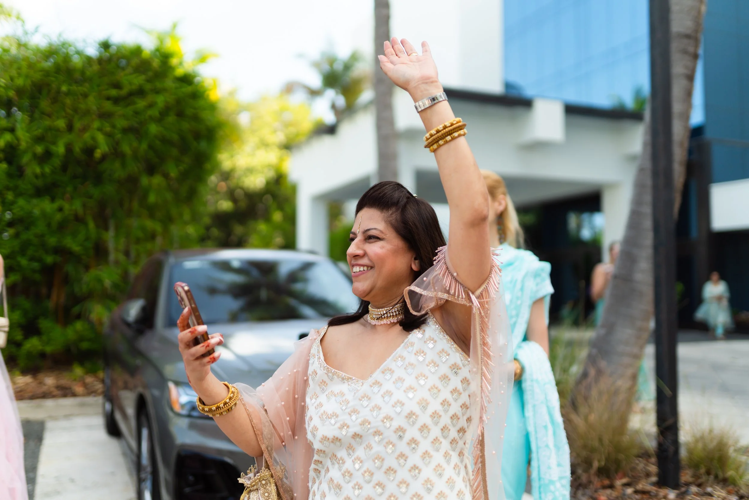 A woman in traditional Indian attire smiling and waving while holding a smartphone at a festive outdoor event.