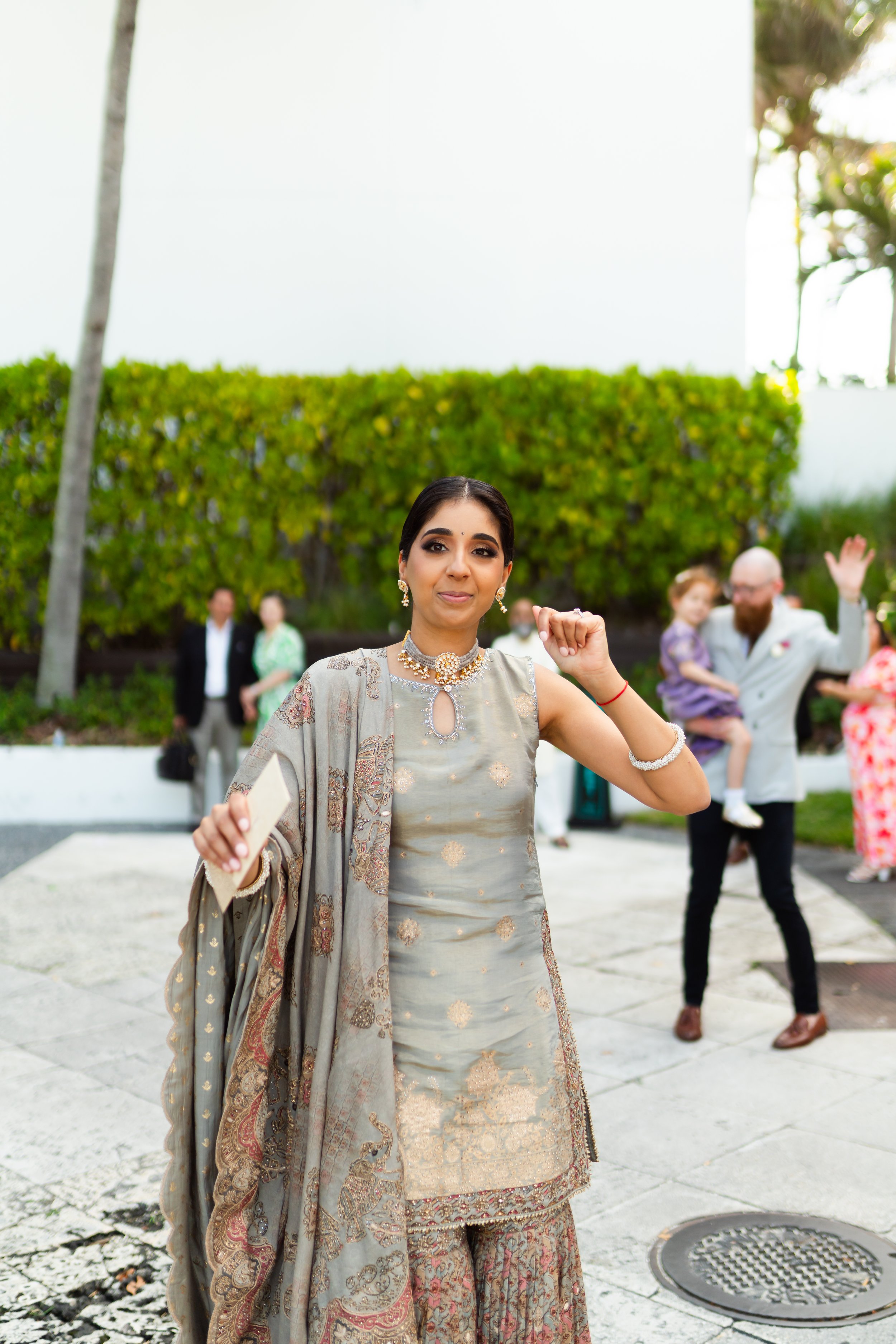 A woman in traditional Indian attire, holding a phone, standing outdoors with a group of people in the background.
