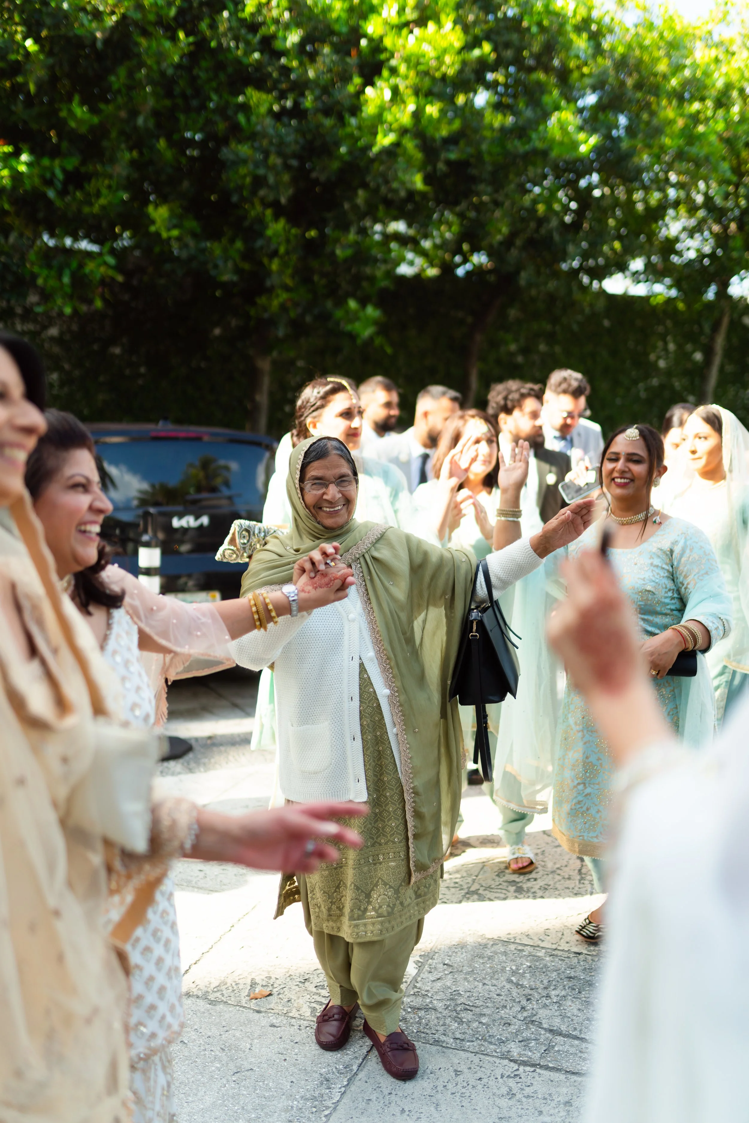 A group of people, mostly women dressed in traditional South Asian attire, celebrating outdoors with smiling and dancing, surrounded by green trees.