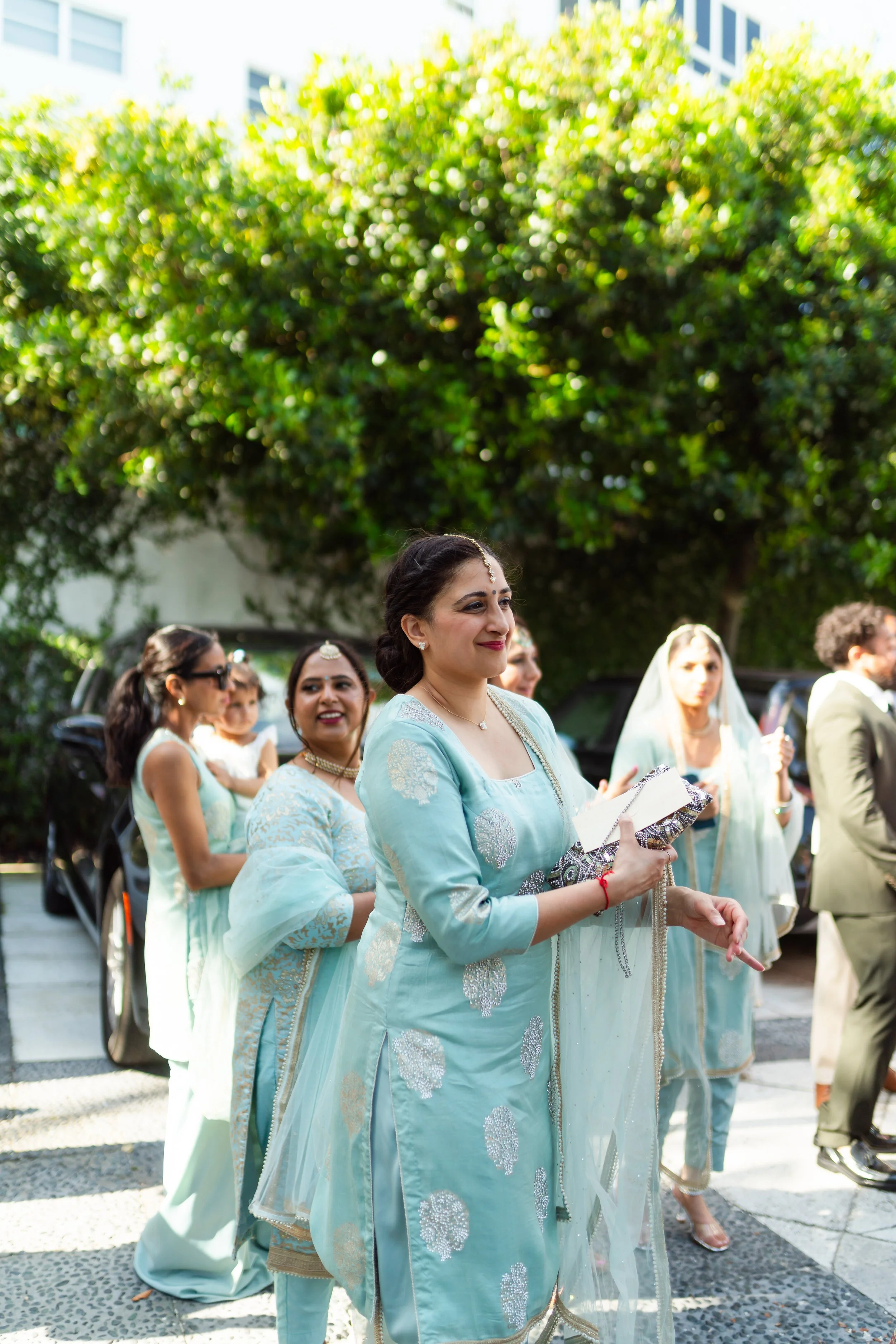 Group of women in traditional Indian attire standing outdoors during daytime, with greenery and buildings in the background.
