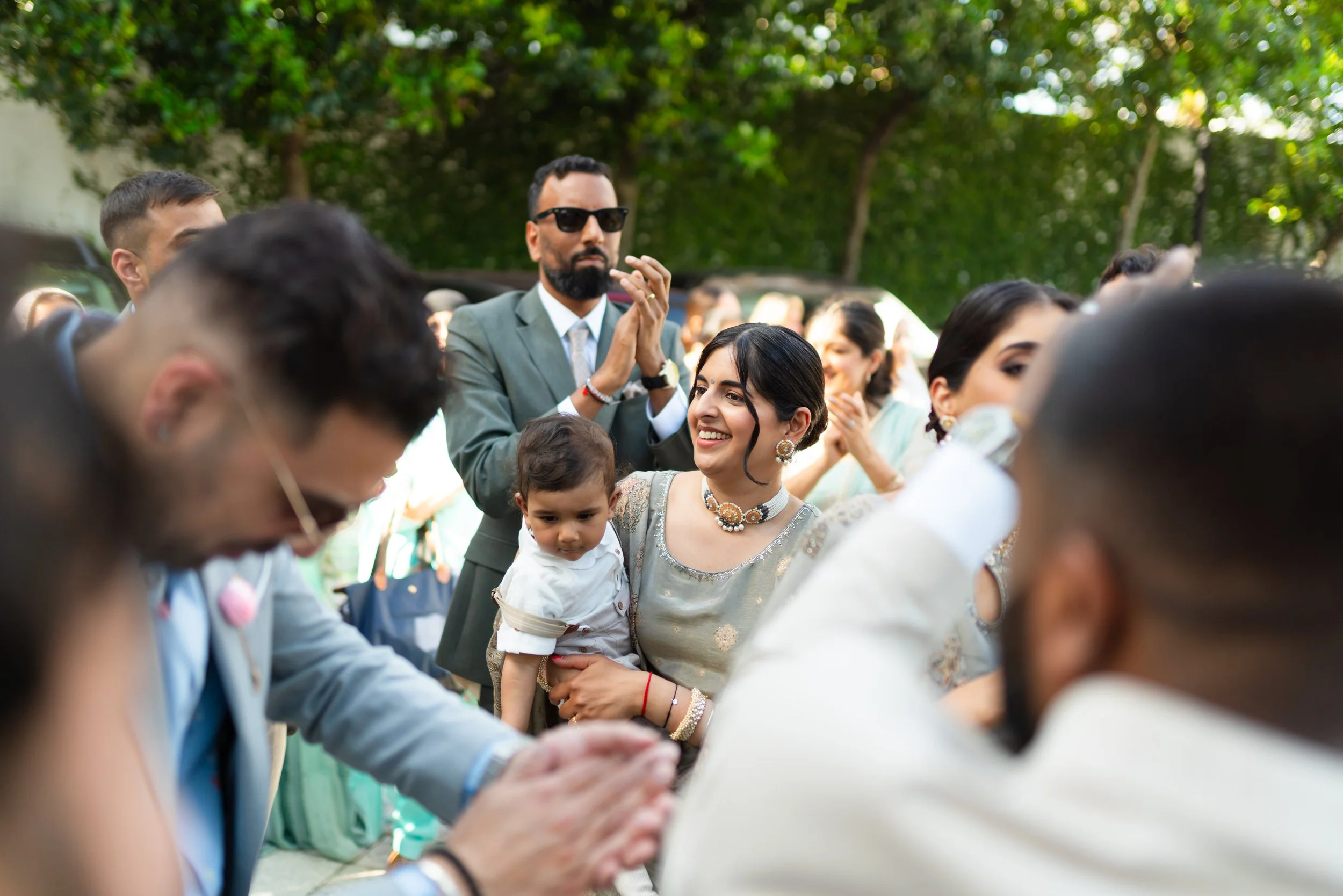 Group of people at an outdoor gathering, with a woman holding a young child, smiling, surrounded by others clapping and engaging in celebration.
