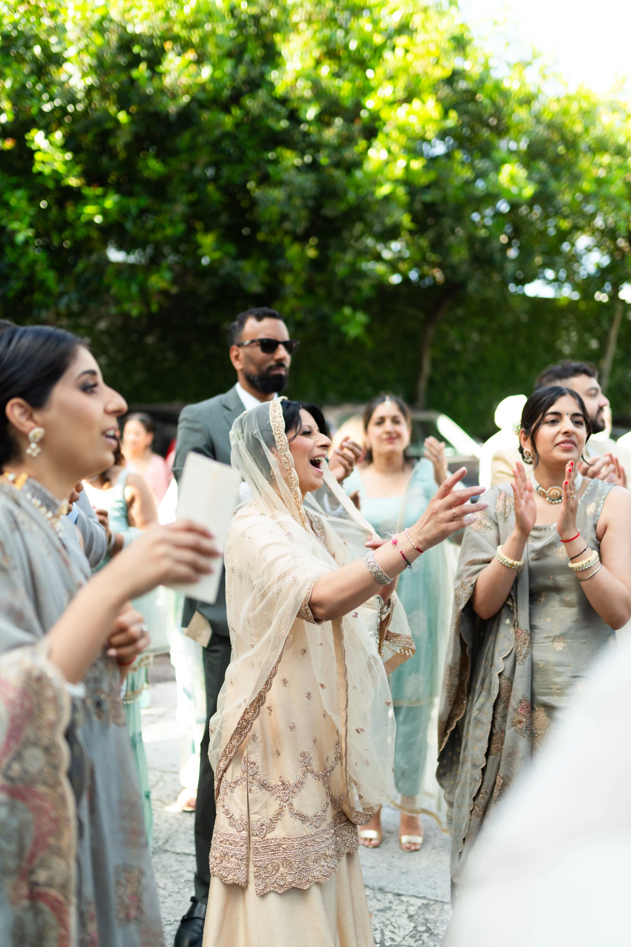 Group of people dressed in traditional South Asian attire enjoying an outdoor celebration, some clapping and smiling.