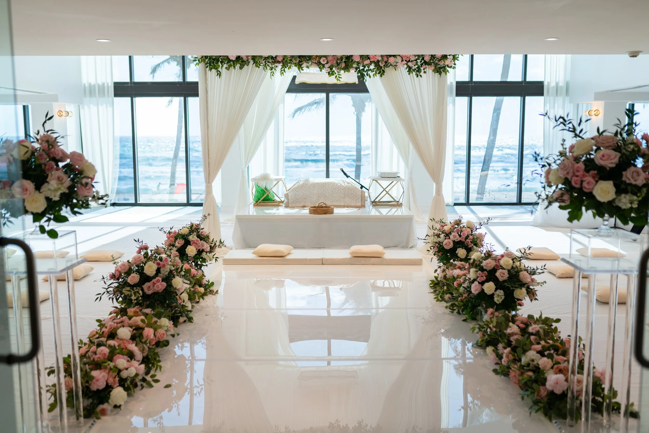 Wedding altar decorated with pink and white flowers, set up in a bright room with large windows and ocean view