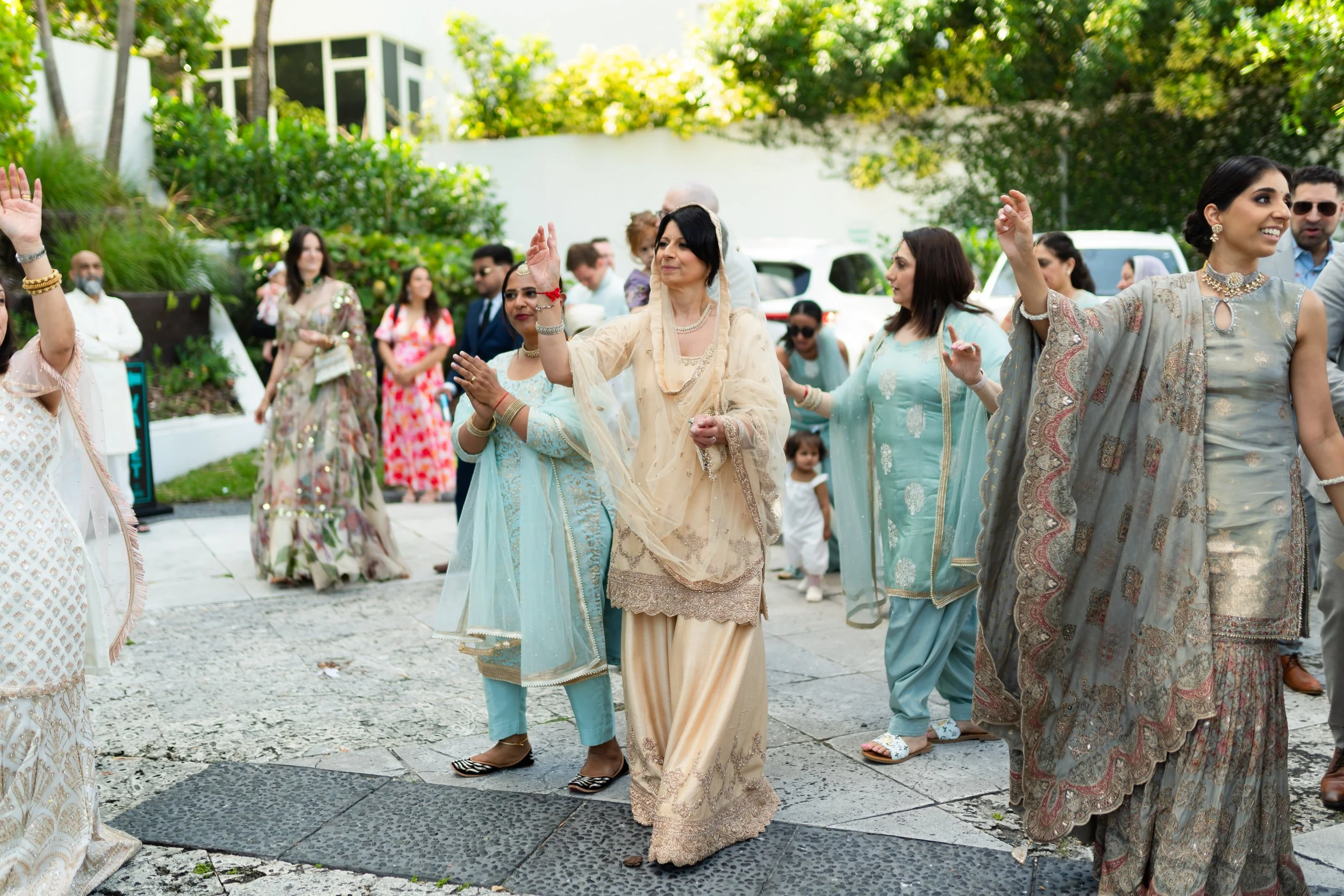 Group of people in traditional South Asian attire dancing outdoors on a stone-paved area surrounded by greenery during a celebration.