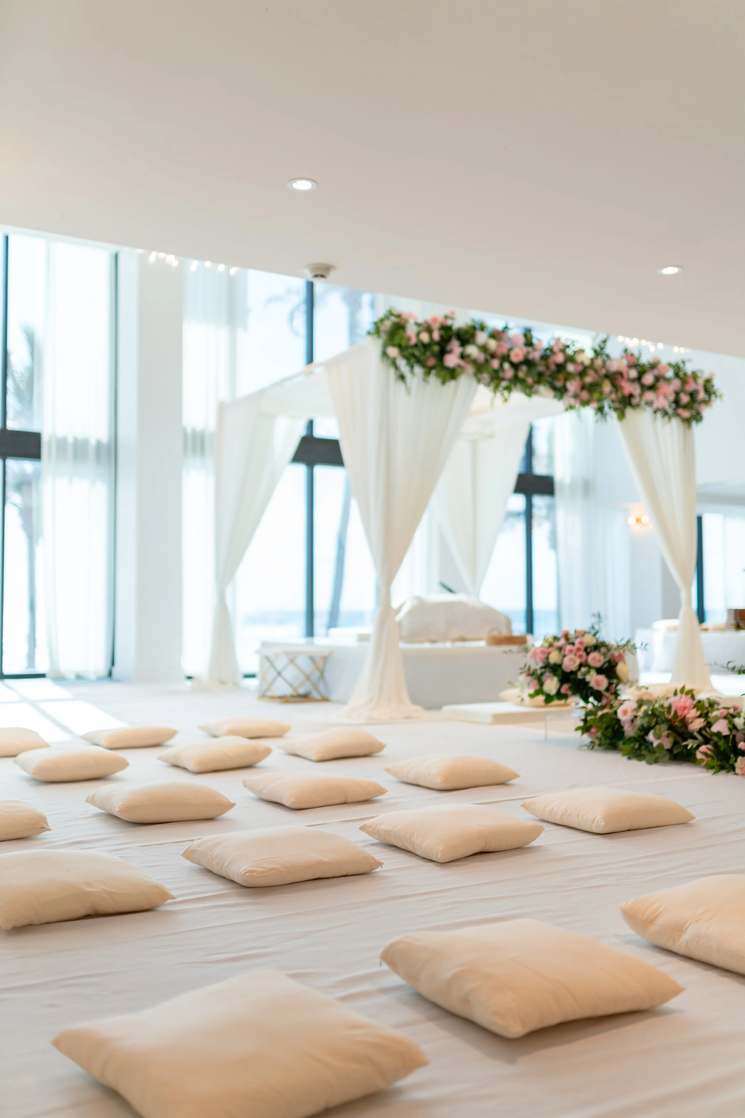 Indoor wedding setup with a floral canopy and cushions on the floor for seating, bright natural light from large windows.