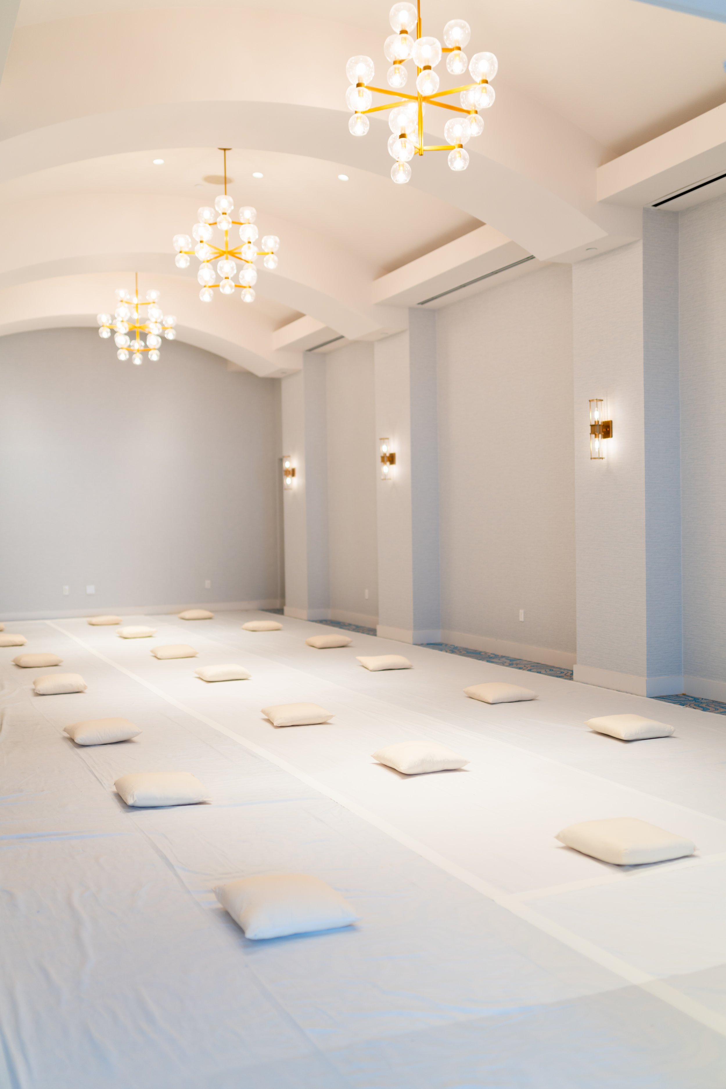 A spacious, white event room with multiple modern chandeliers hanging from the ceiling and wall-mounted light fixtures. The floor is covered with protective white sheets, and there are evenly spaced white pillows placed on the floor, likely for a med