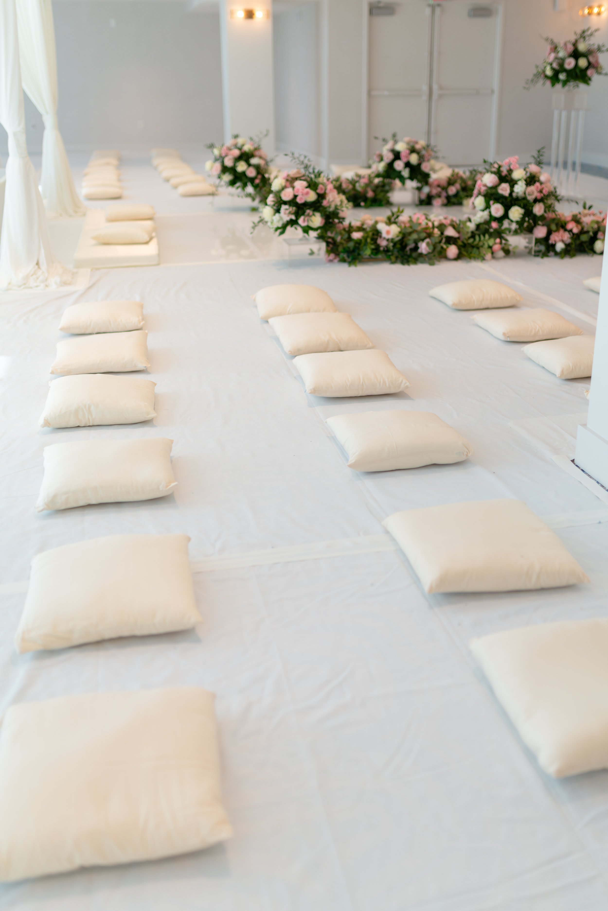 Indoor wedding or event space decorated with pink and white flowers, with cushions arranged on a white floor for seating.