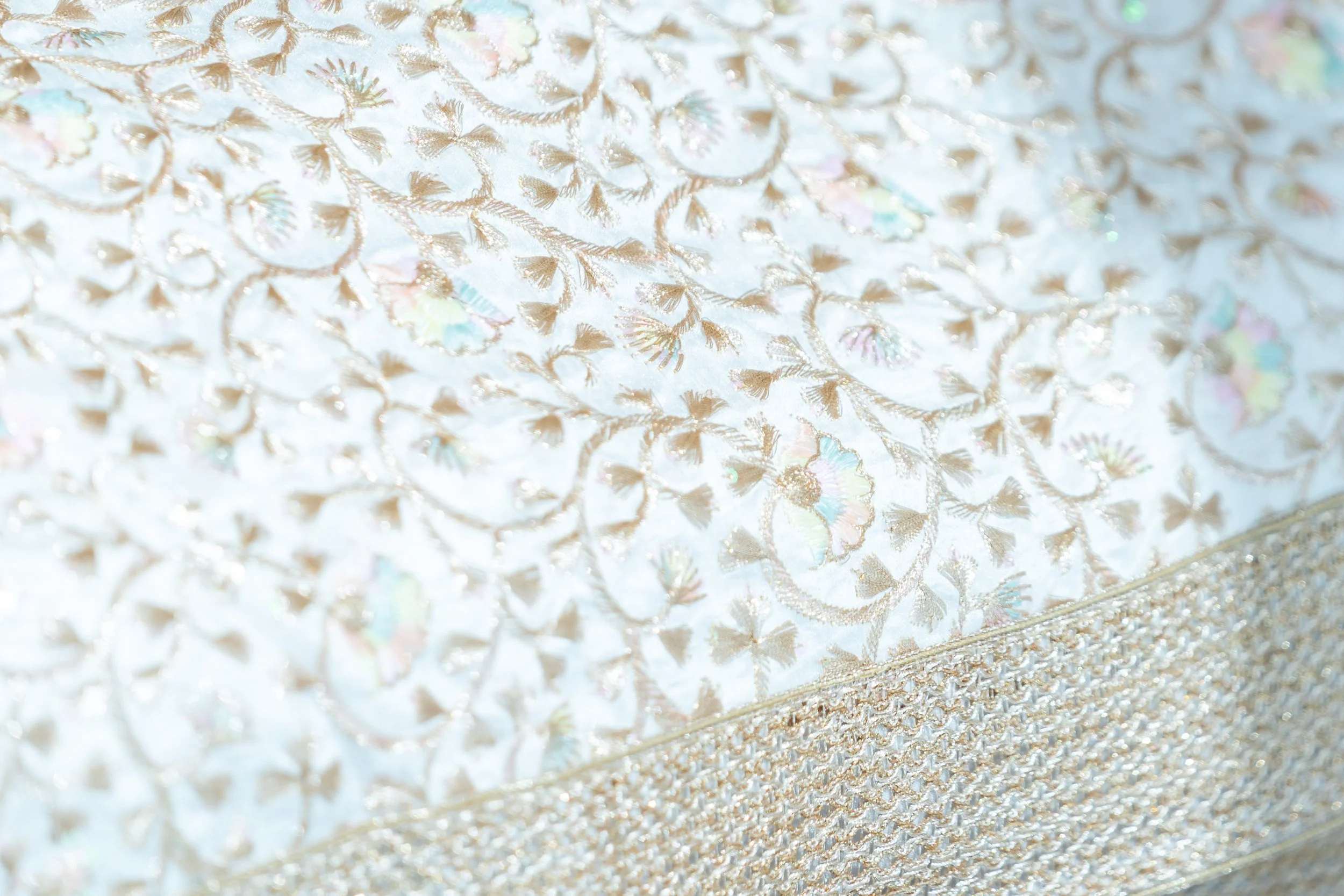 Close-up of an ornate fabric with intricate floral embroidery and shimmering decorations in light pastel colors.