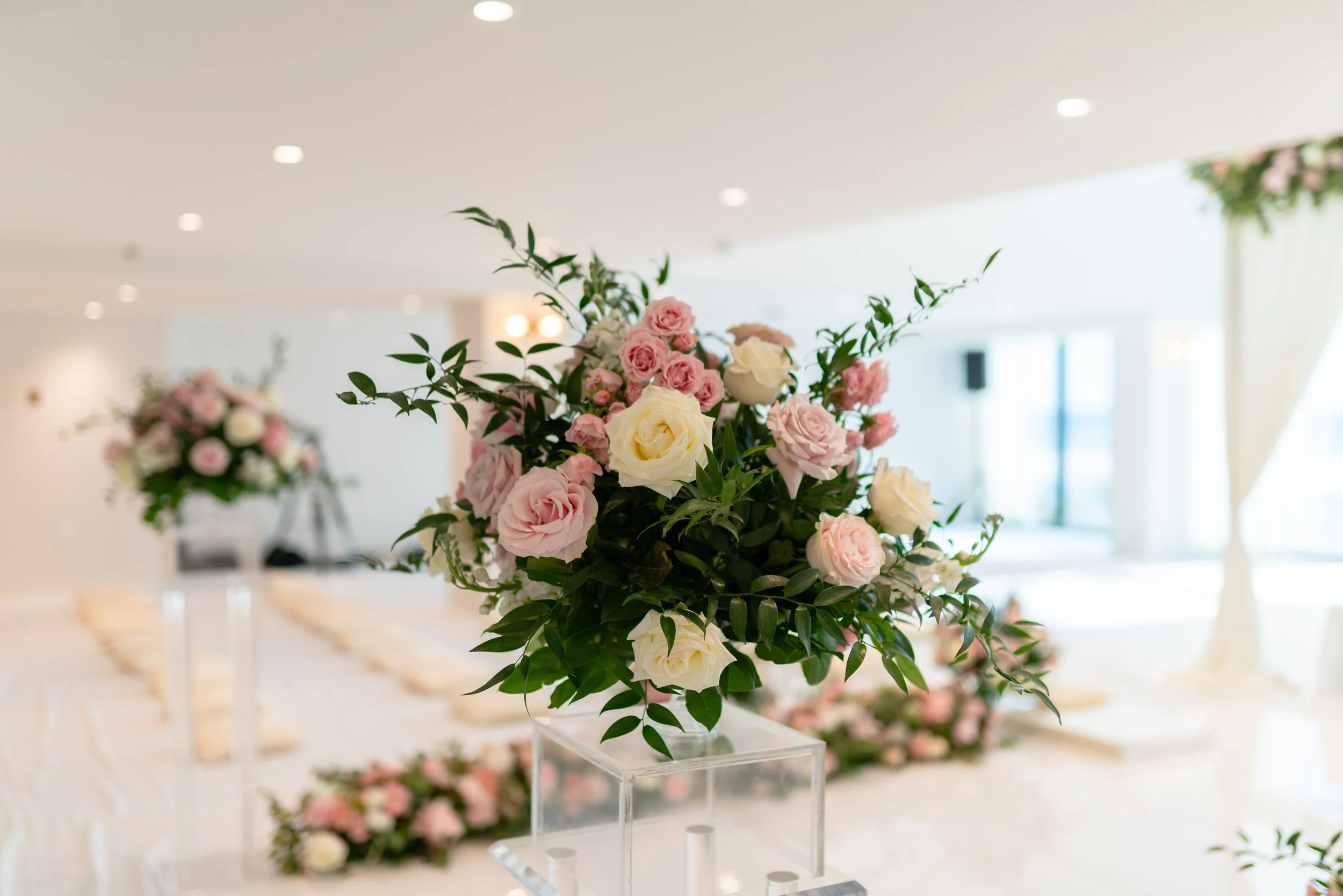 Arrangement of pink and white roses with green foliage on a transparent stand in a bright indoor setting, likely for a wedding or event.