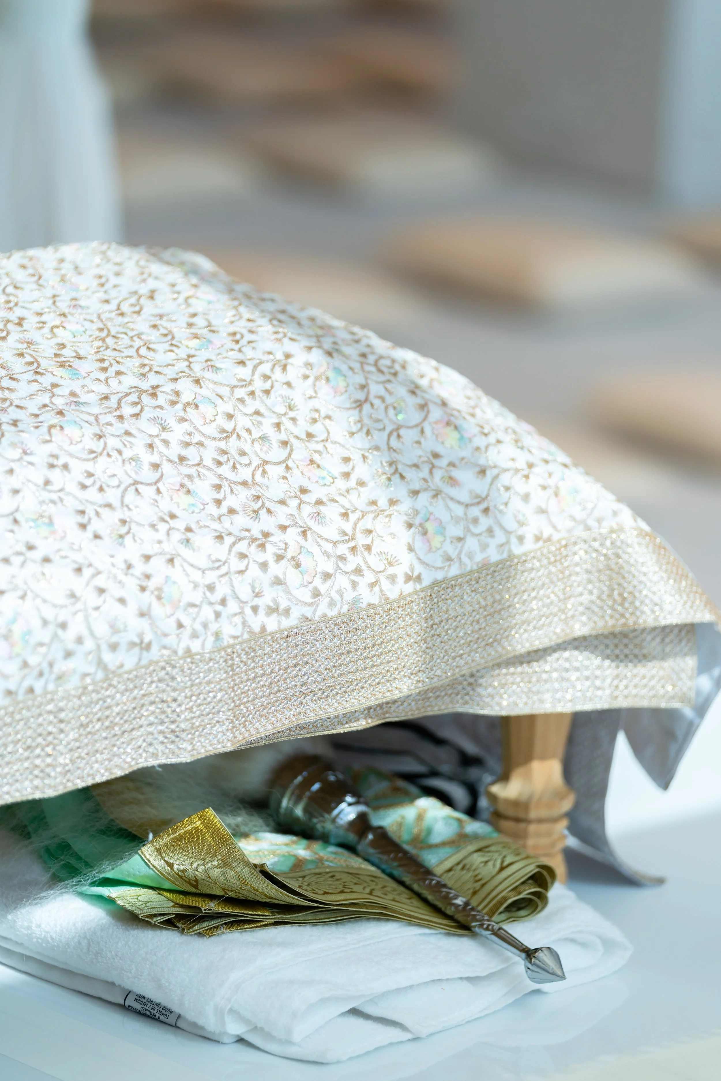A decorative cake with white icing and gold accents, covered with a lace overlay, with religious items beneath including a spoon and fabric, all on a white table.