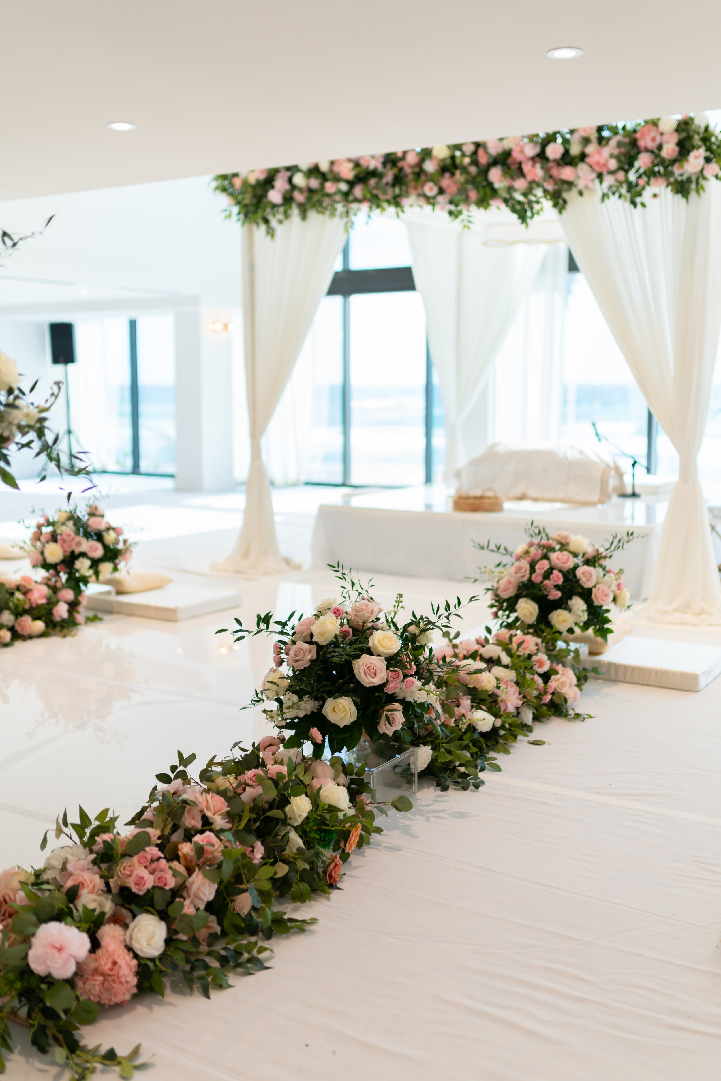 Indoor wedding ceremony setup with floral arch and flower arrangements in pink and white.