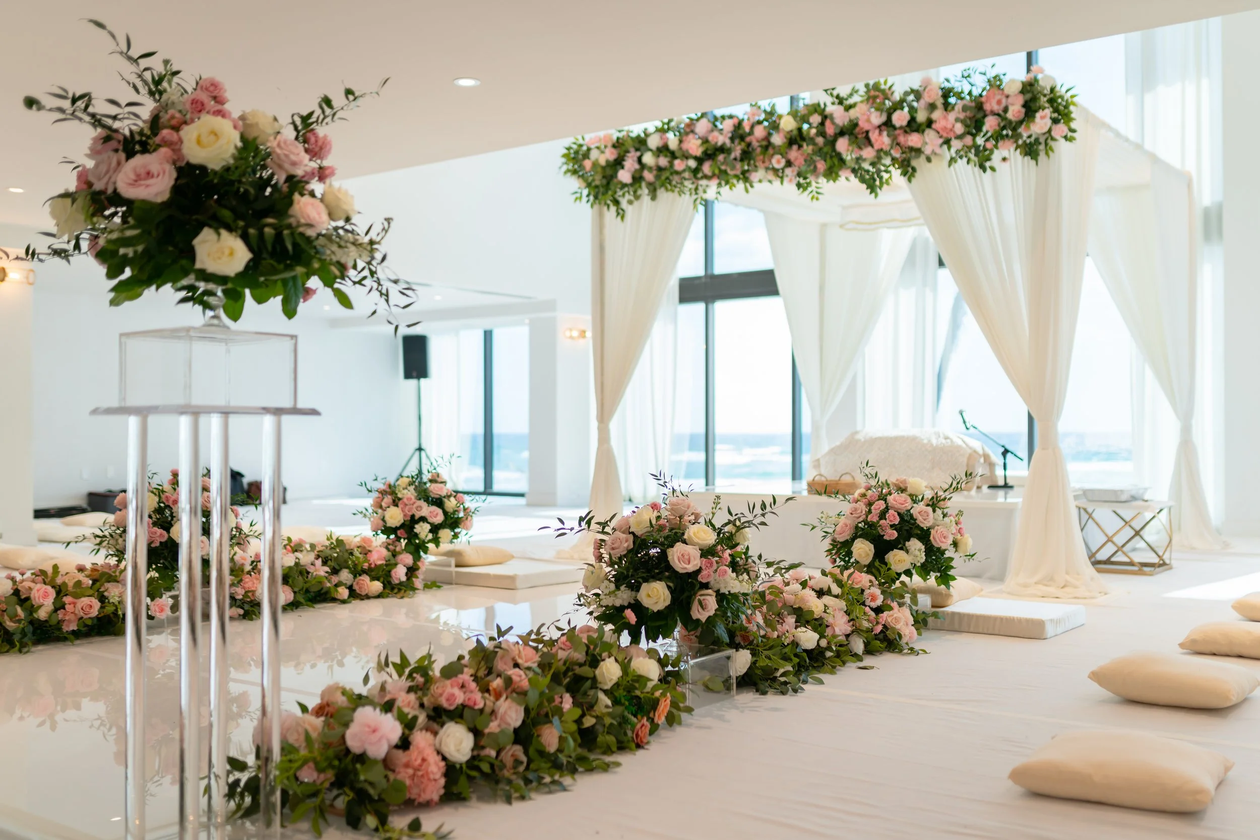 Elegant wedding ceremony setup with a floral arch, pink and white roses, and soft white cushions on the floor, with large windows and flowing white curtains in the background.