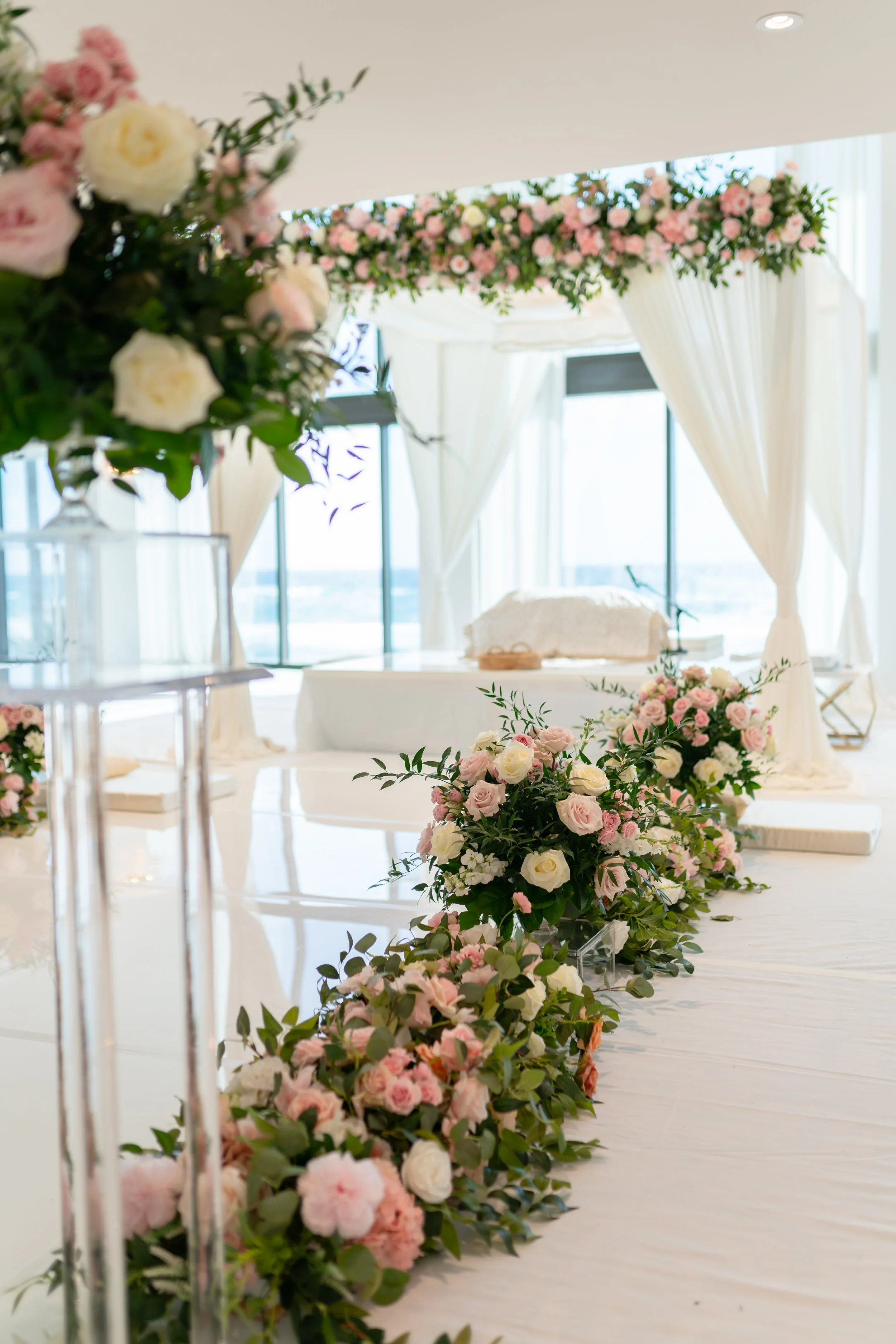 Elegant indoor wedding ceremony setup with a flower-lined aisle, white drapes, large windows, and a decorated arch with pink and white flowers.