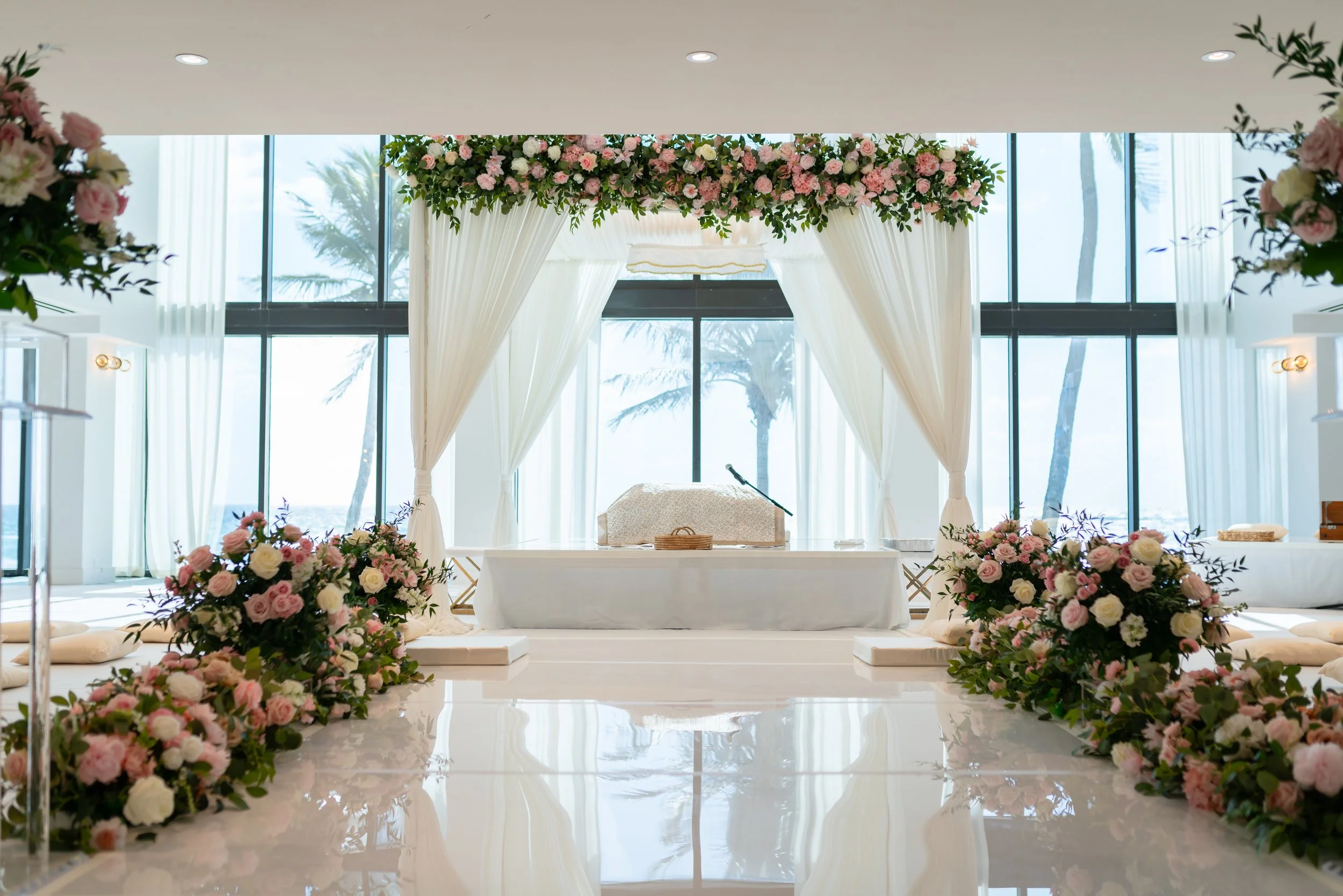Wedding altar decorated with pink and white flowers, white curtains, and palm trees visible through large glass windows.