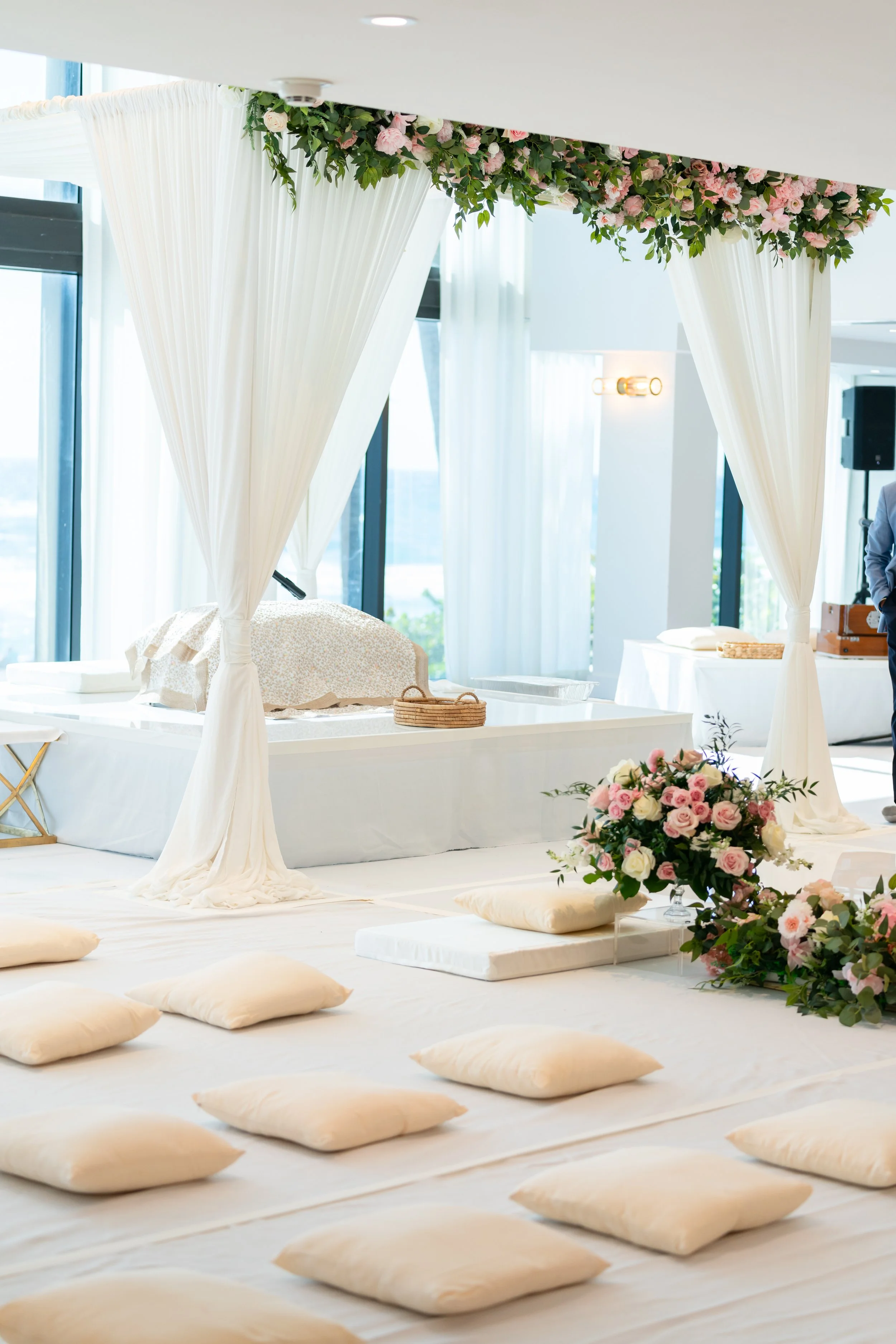 Wedding ceremony setup with flower arch, cushions on the floor, and a decorated altar in a bright, modern space with large windows.