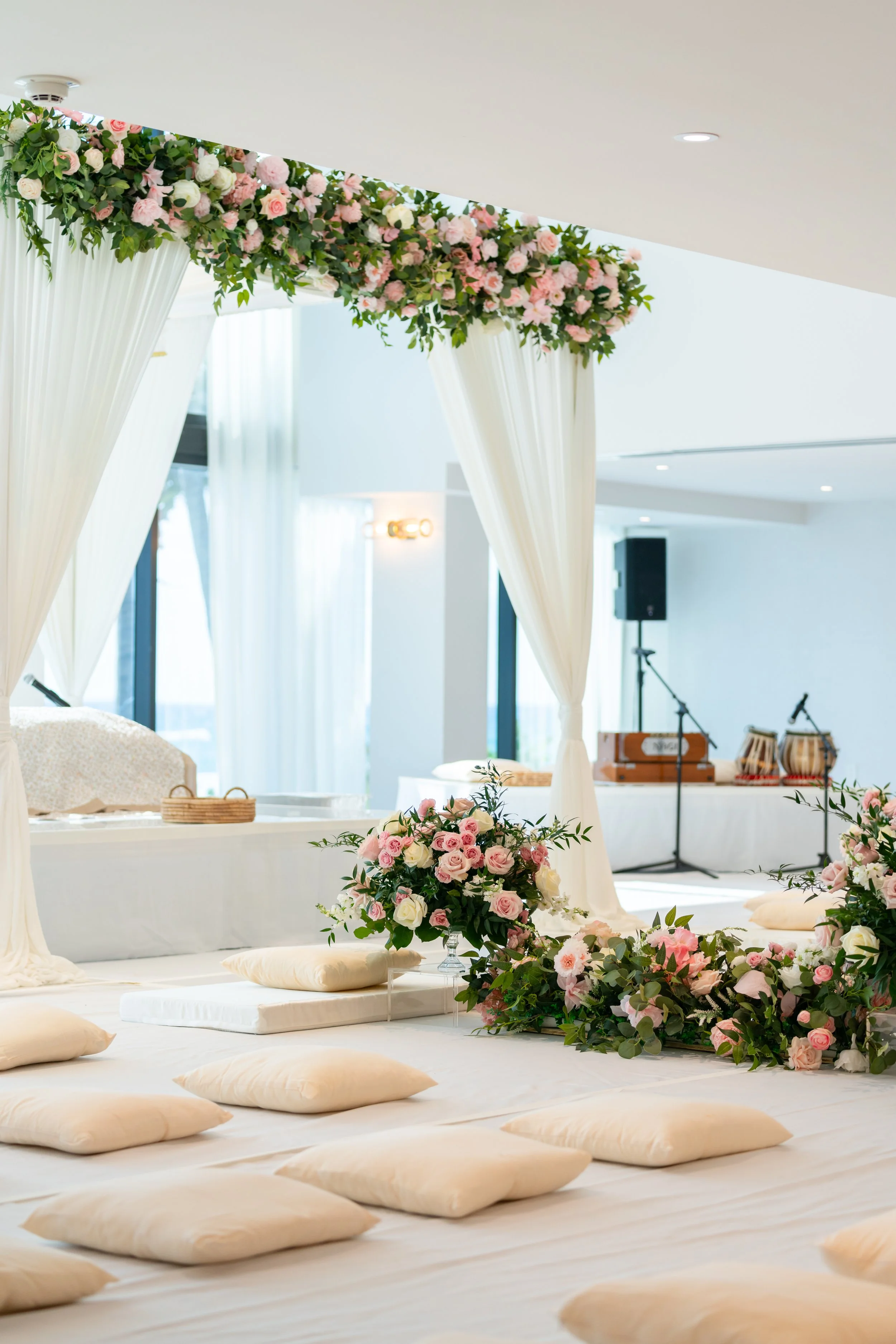 Elegant indoor wedding setup with white draped curtains and pink and white floral arrangements on the floor and above the curtains, with cushions and soft lighting.