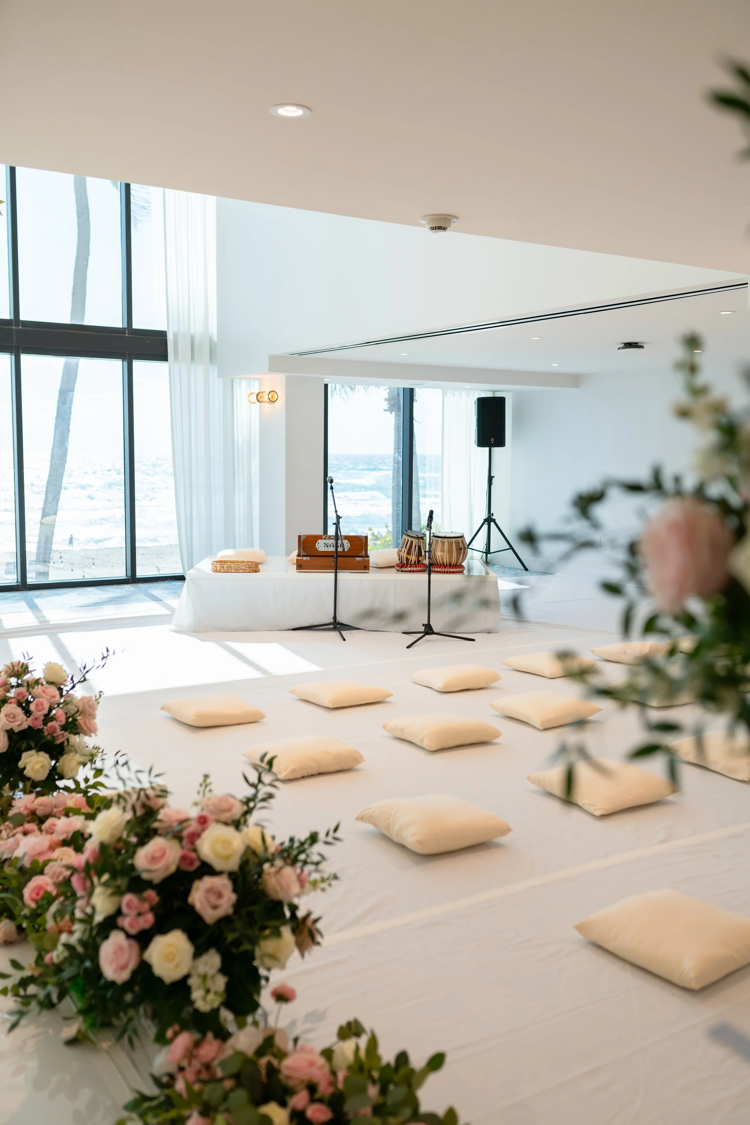Indoor space set up for a musical performance with cushions on the floor, a table with drums and microphones, and a view of the ocean through large windows.