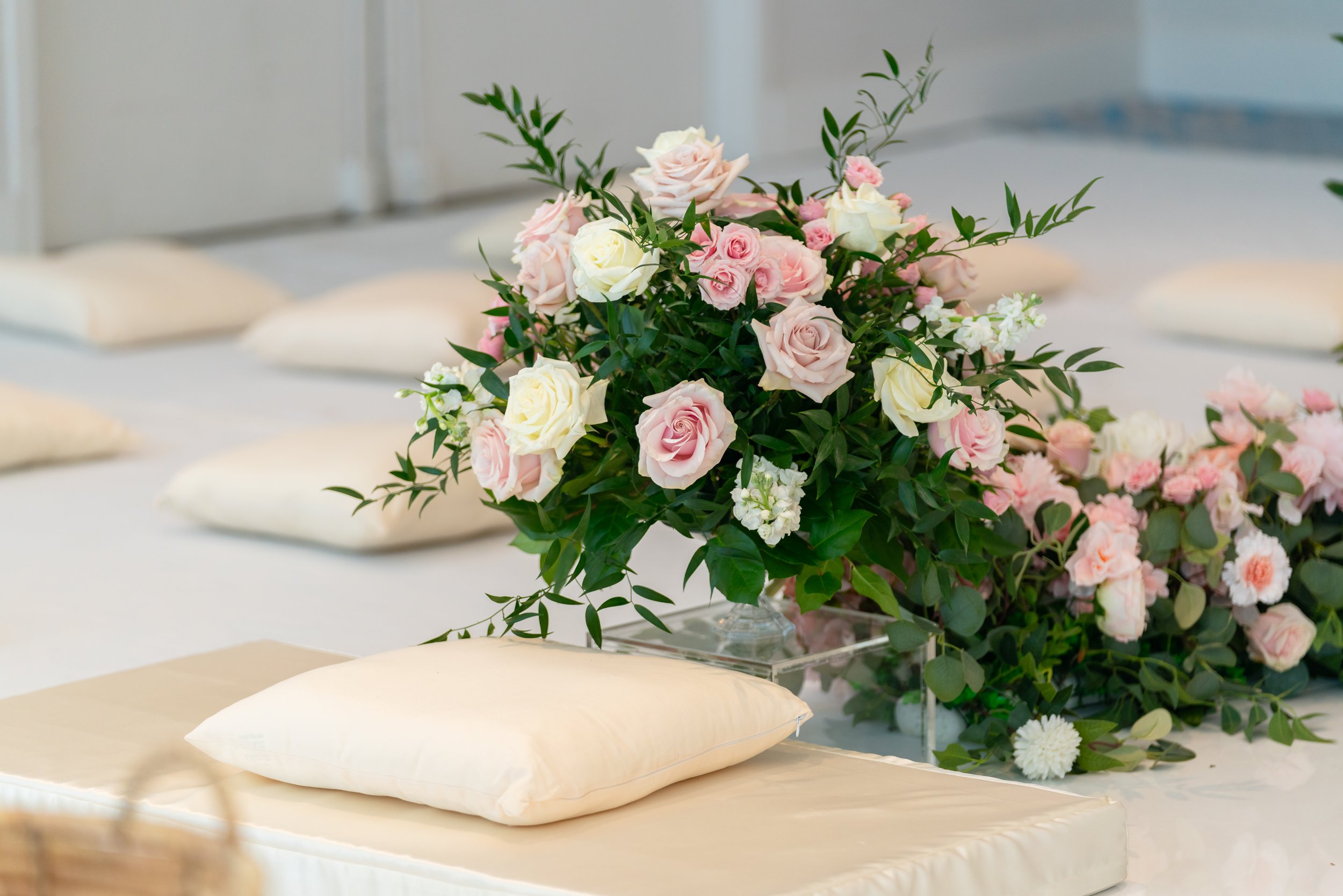 A floral arrangement with pink and white roses and greenery on a white table, with beige pillows in the background.