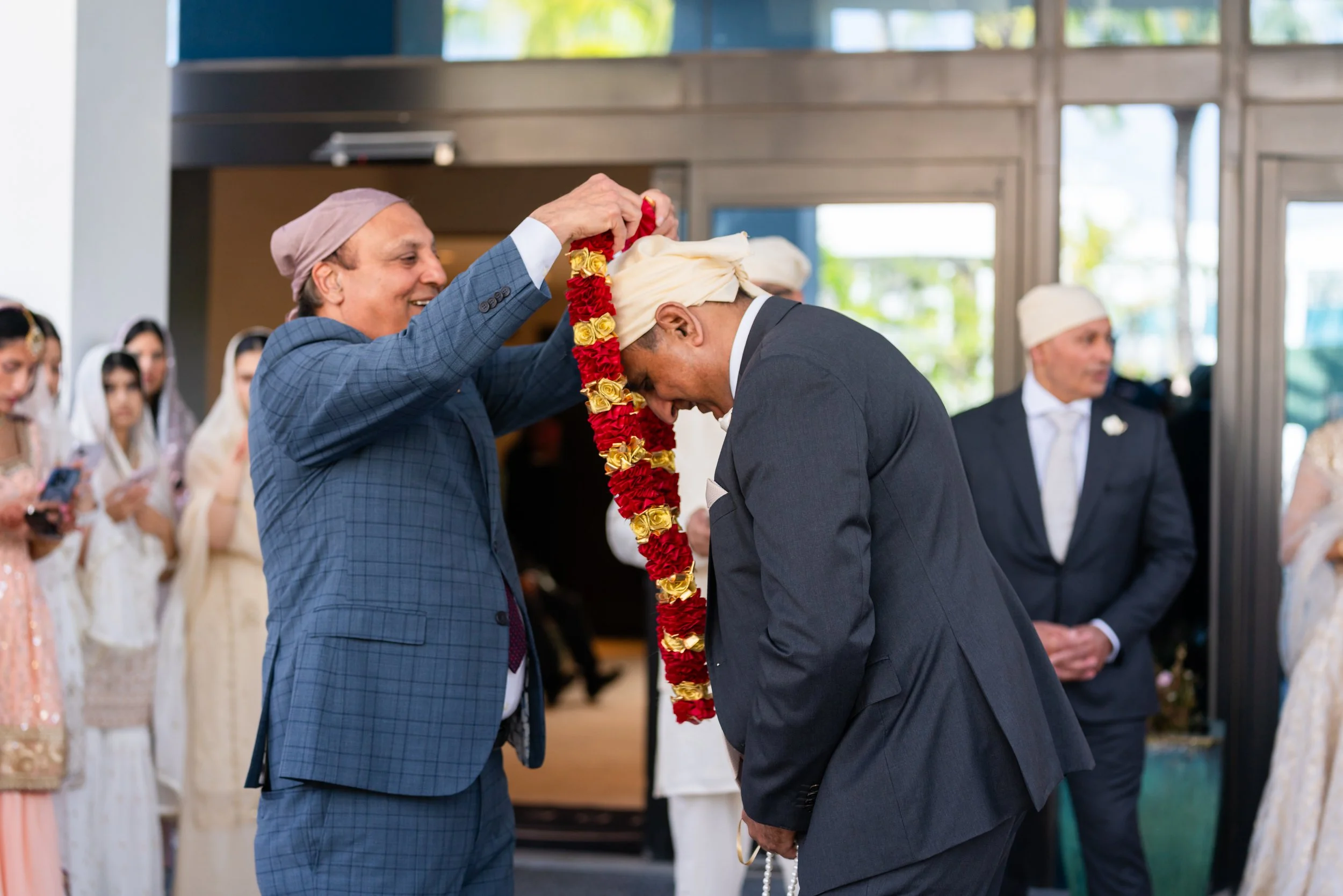 A man in a blue checked suit and pink head covering places a garland of flowers around the neck of another man in a dark suit and white turban, who is bowing his head in respect in a formal indoor setting with several people dressed in traditional at