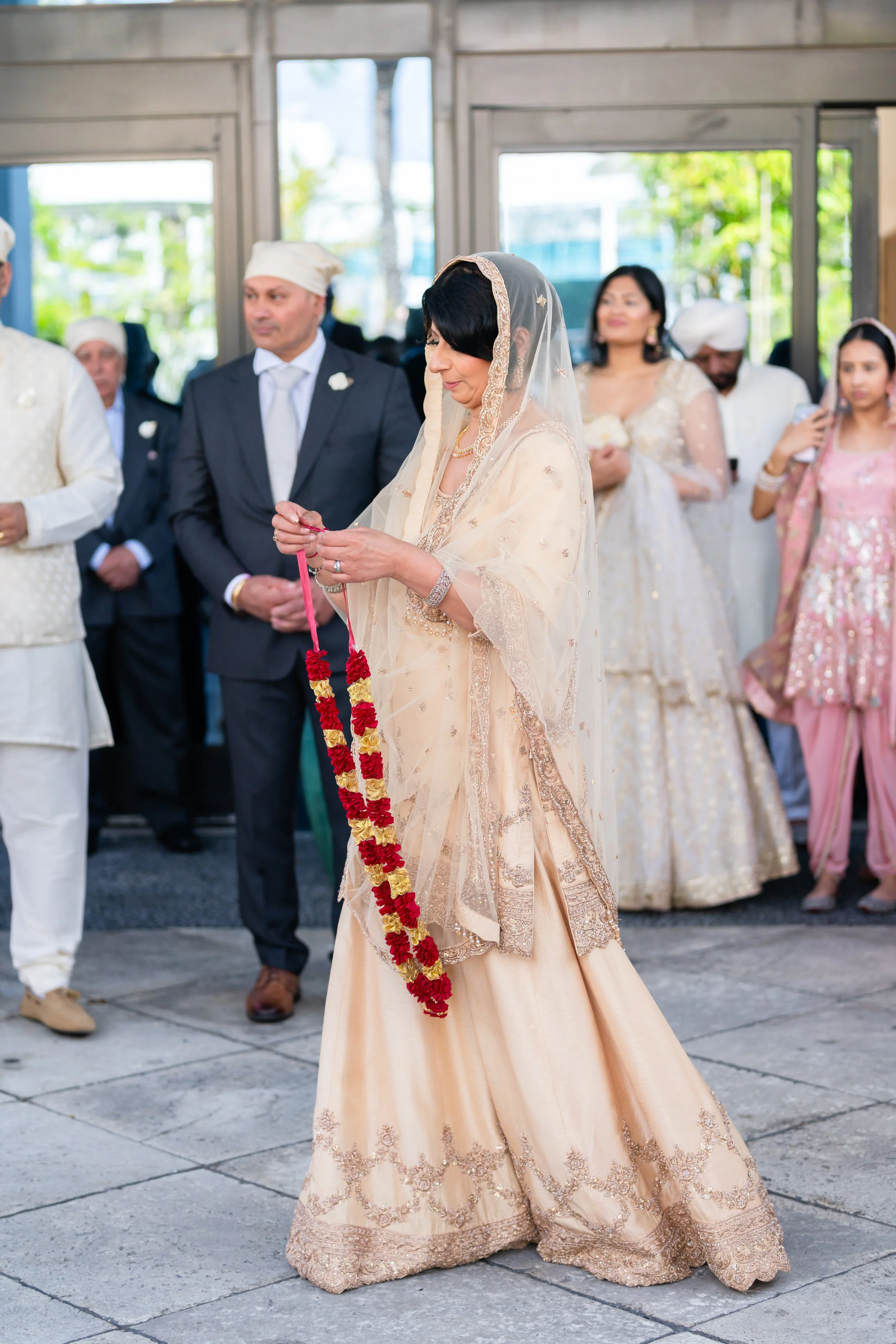 A woman in traditional South Asian attire holds a flower garland during a wedding ceremony, surrounded by other guests.