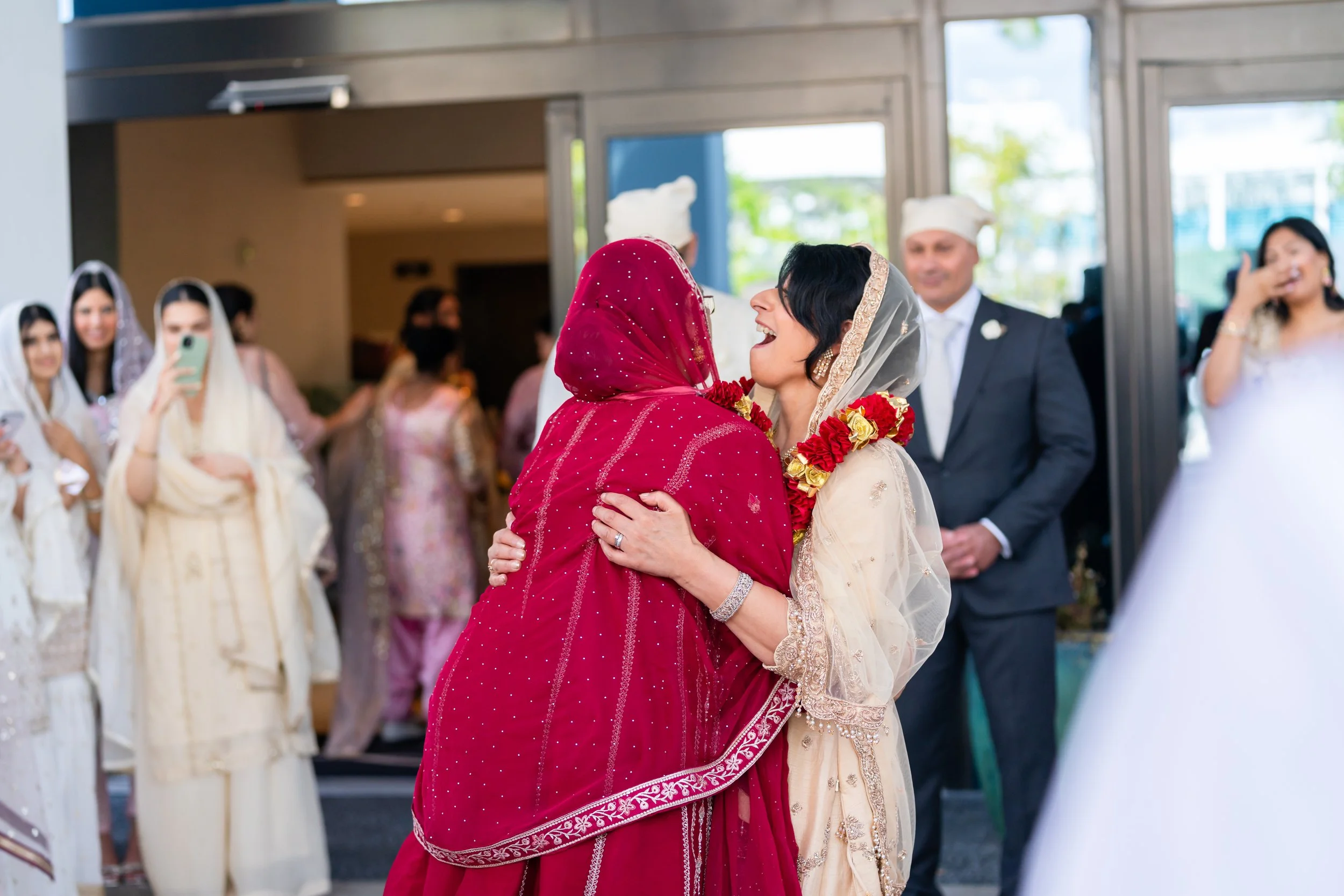 Two women hugging at a wedding celebration, one in a red traditional dress and the other in a light-colored dress with a floral garland on her neck, surrounded by wedding guests in traditional and modern attire.