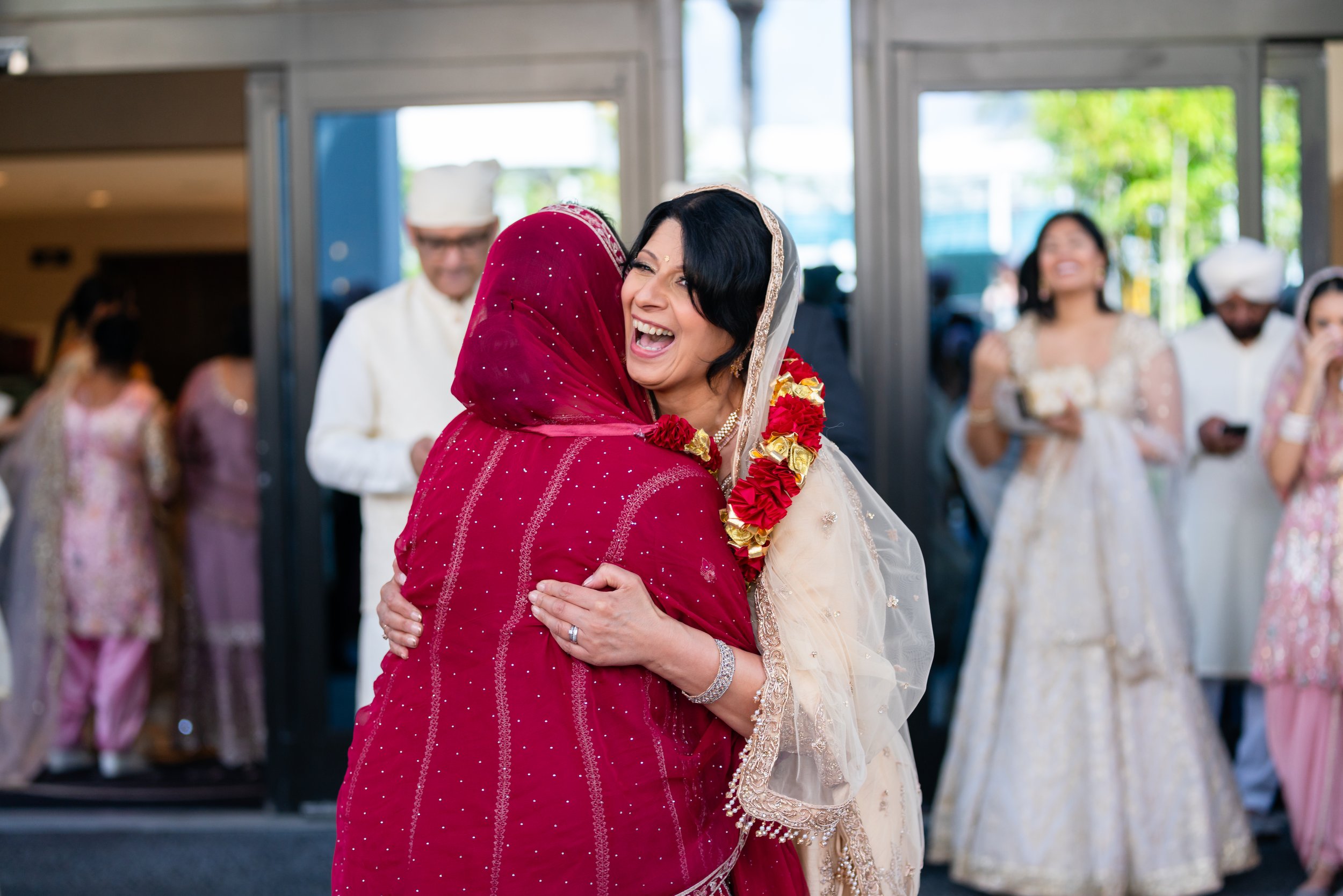 Two women embracing at a wedding, one wearing a red sari and the other in traditional white attire with a floral garland around her neck, smiling and laughing. In the background, other guests are seen standing and chatting.