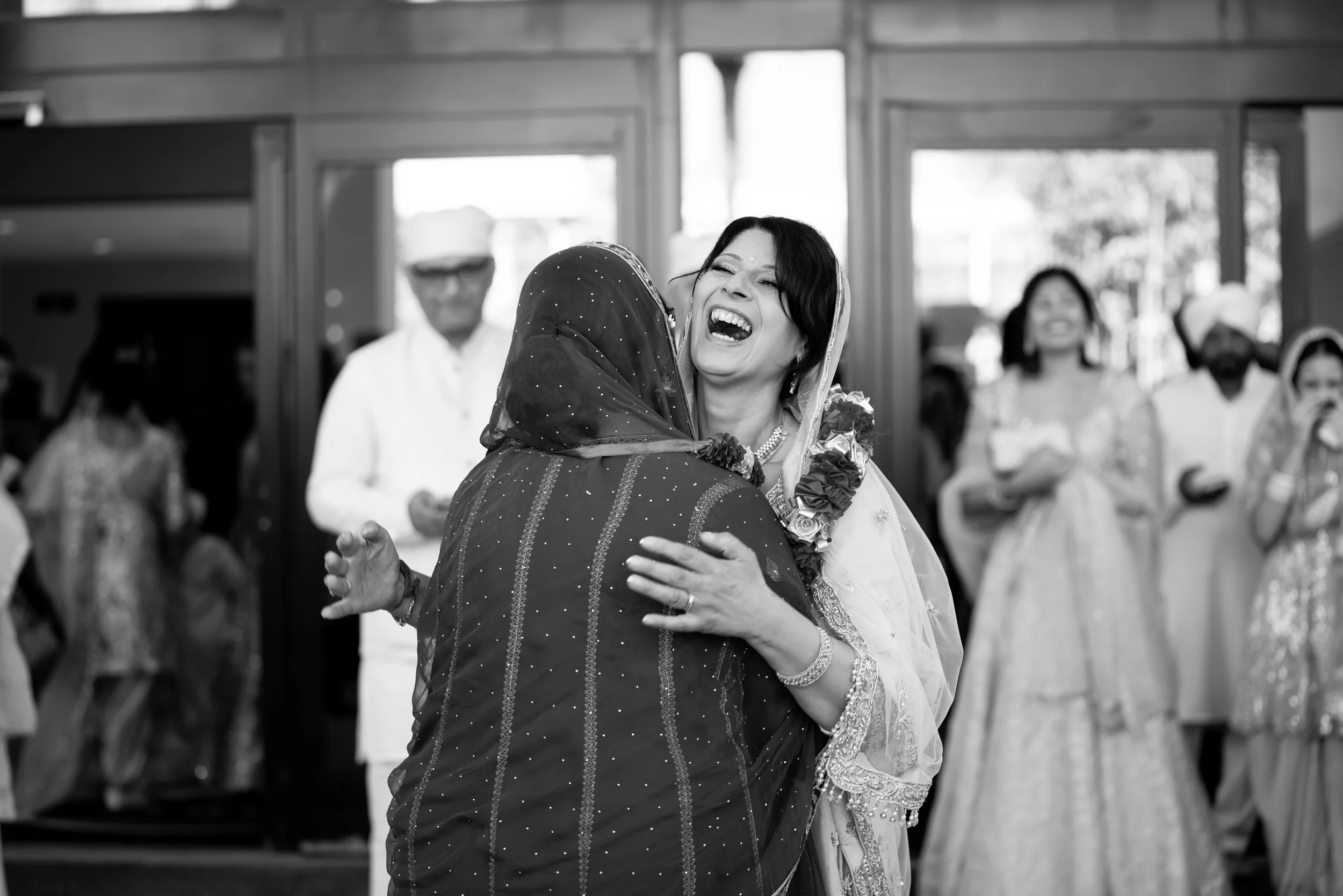 Two women embracing at a celebration, laughing happily, with guests in traditional attire standing behind them.