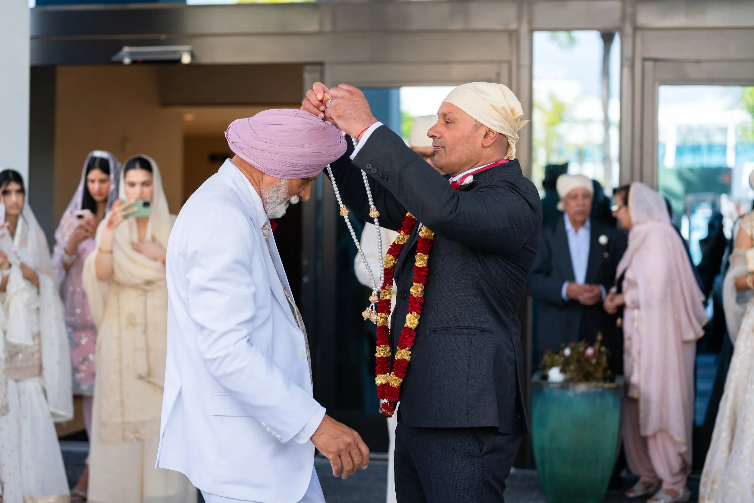 A man in a suit and garland places a beaded necklace around the neck of an elderly man in a white suit and pink turban during a celebratory event.