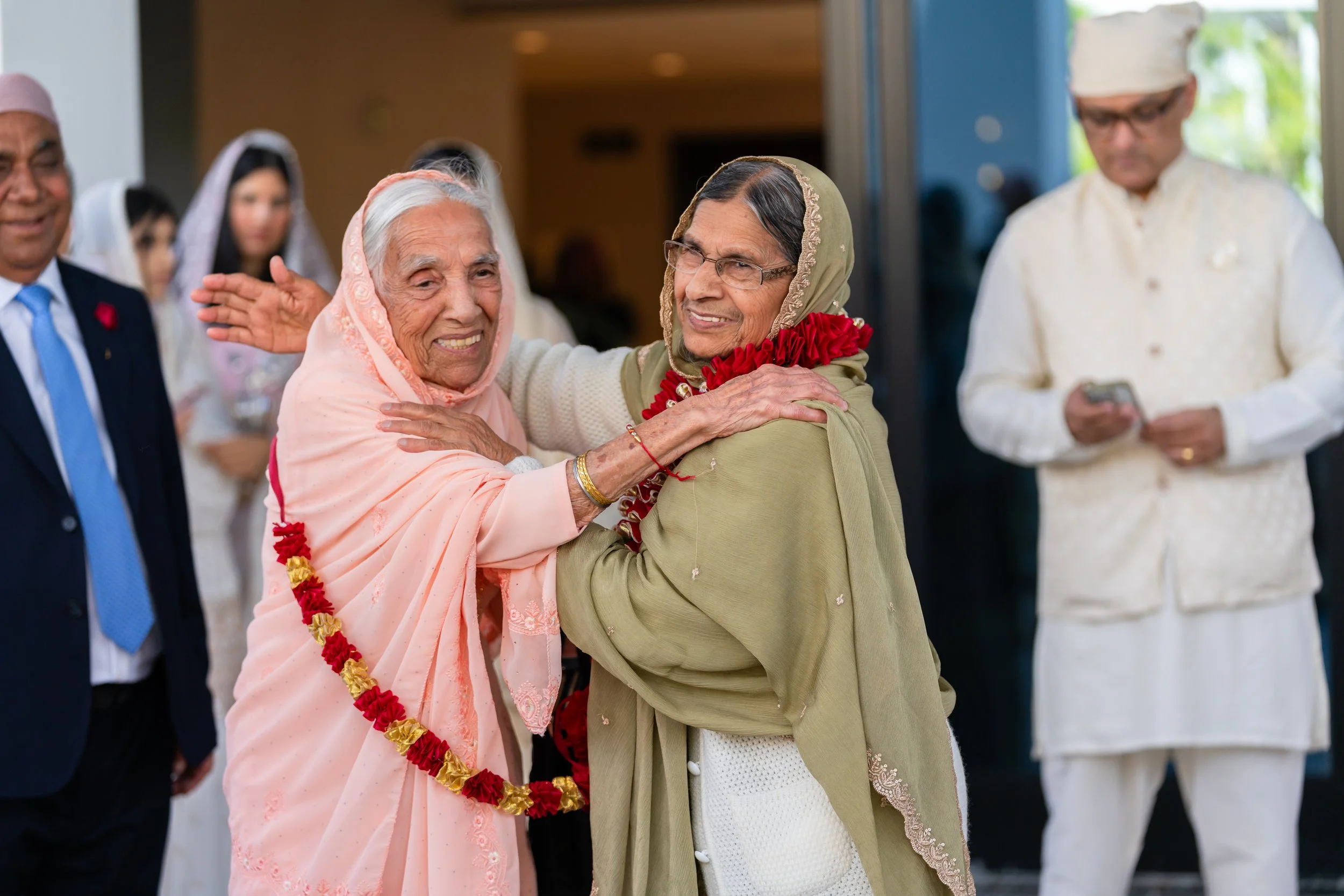Two elderly women in traditional South Asian attire hugging each other at a celebration, with a group of people in the background.