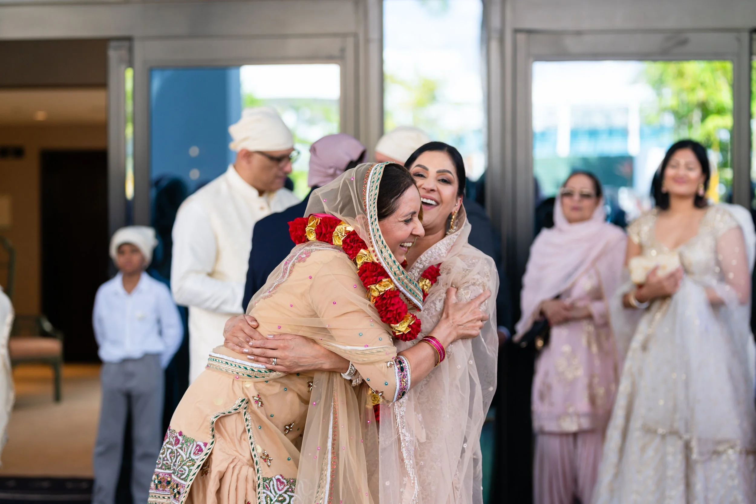 Two women, one in traditional South Asian attire with a flower garland, hugging and smiling at a celebration. Several other people are smiling and standing in the background.