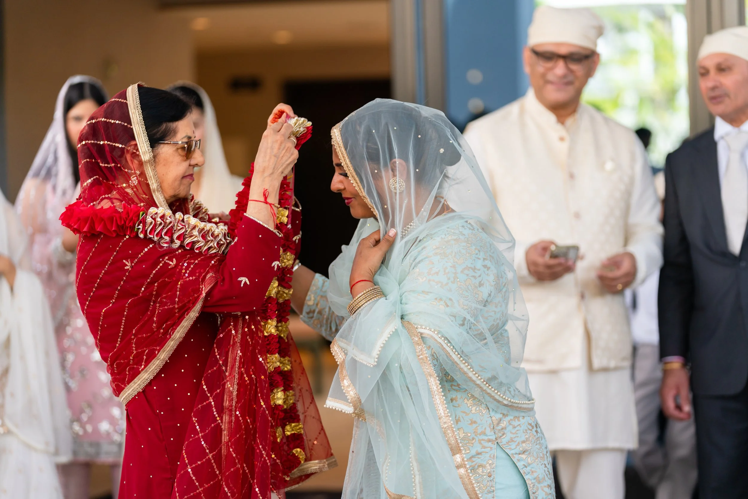 Indian bride dressed in light blue saree and veil receiving floral garland from an older woman in red traditional attire during a wedding ceremony, with two men and other women in the background.