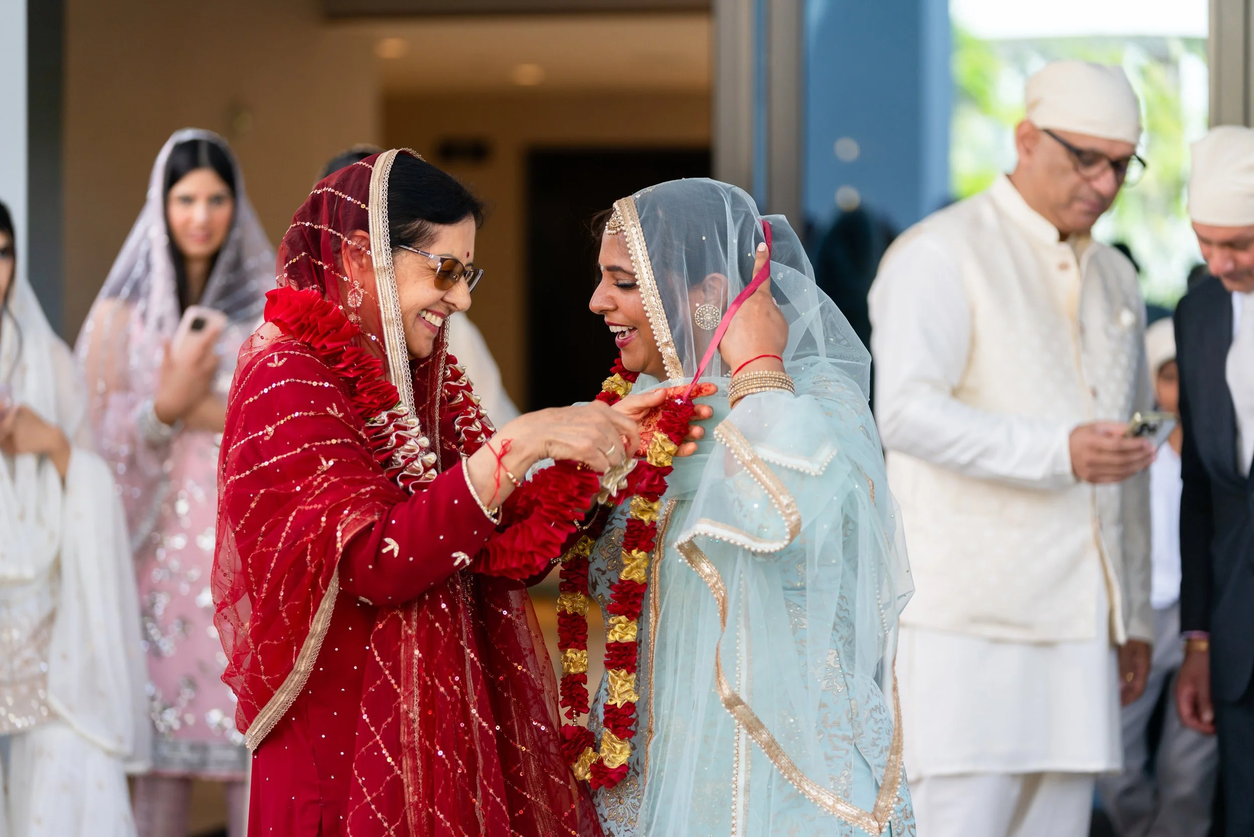 Two women dressed in traditional South Asian attire, one in red and the other in light blue, exchanging garlands at a cultural wedding ceremony. Several other people are in the background, some using their phones.