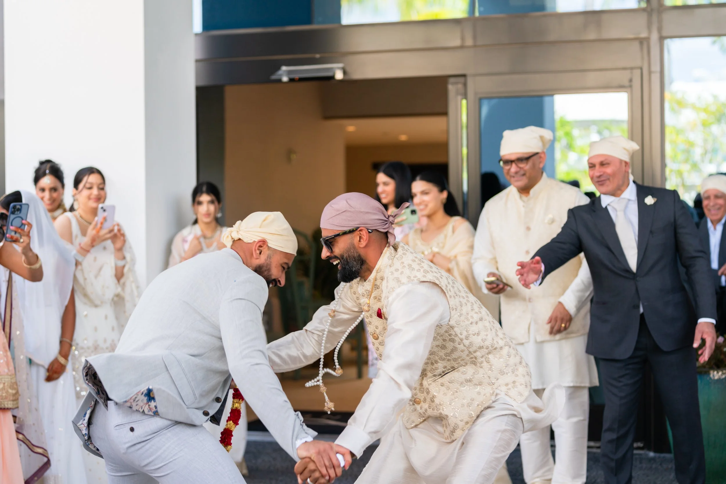 Two men dressed in traditional Indian attire are dancing and holding hands at a wedding celebration, surrounded by friends and family.
