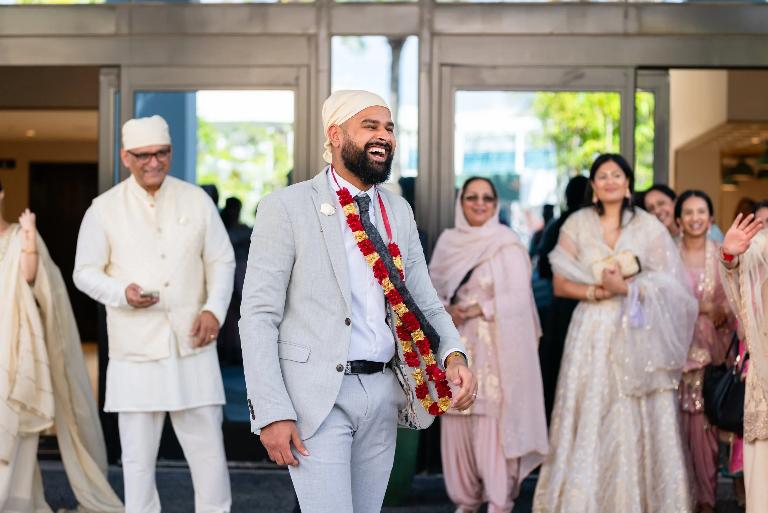 Smiling groom in a light gray suit with a floral garland, surrounded by guests in traditional South Asian attire at a wedding ceremony.