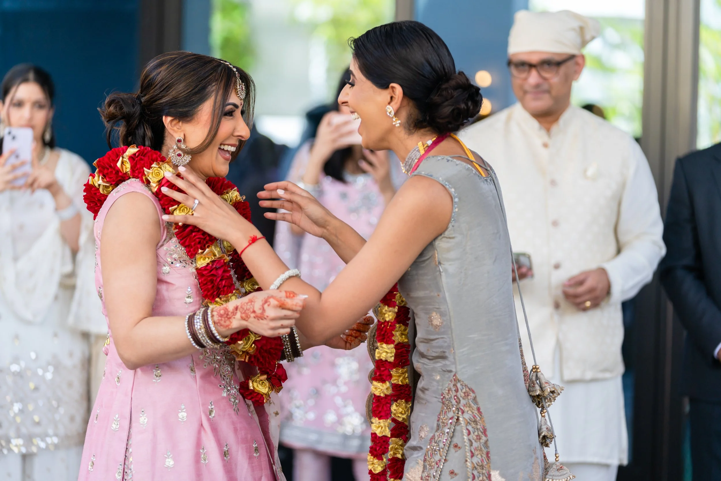 Two women smiling and exchanging hugs at a celebration. The woman on the left is wearing a pink dress with silver embellishments, jewelry, and a floral garland. The woman on the right is dressed in a gray and gold traditional outfit, also wearing a f