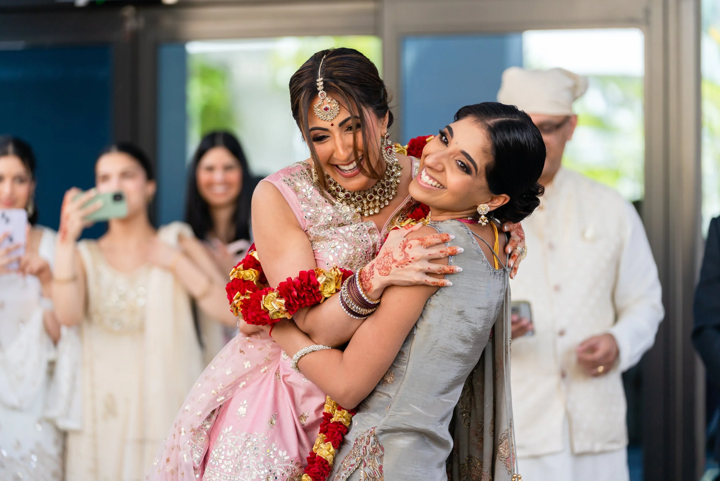 Two women hugging and smiling at a celebration, with one wearing traditional Indian attire and jewelry, and the women in the background taking photos.
