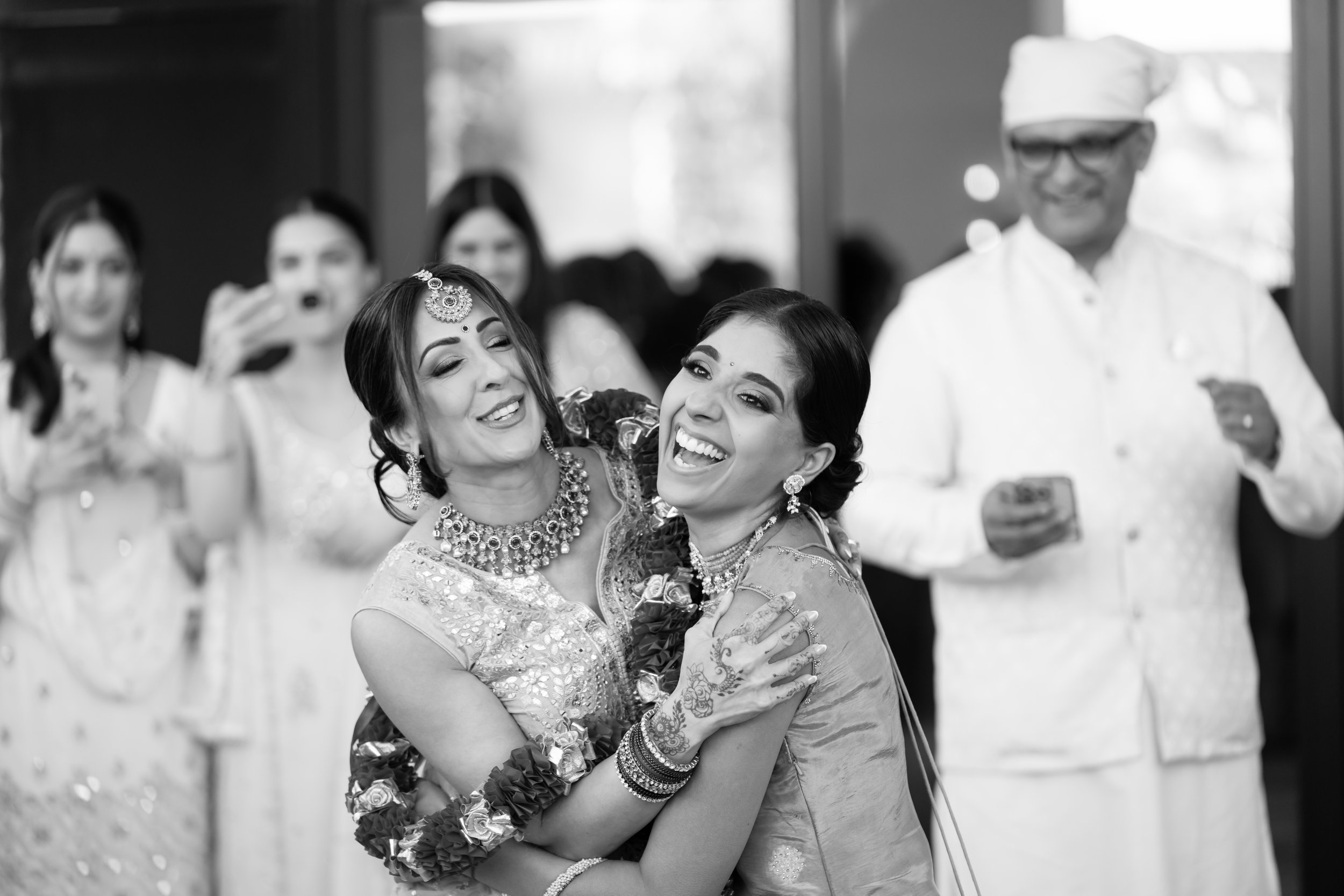 Women dressed in traditional Indian attire and jewelry hugging and smiling, with onlookers in the background at a celebration.