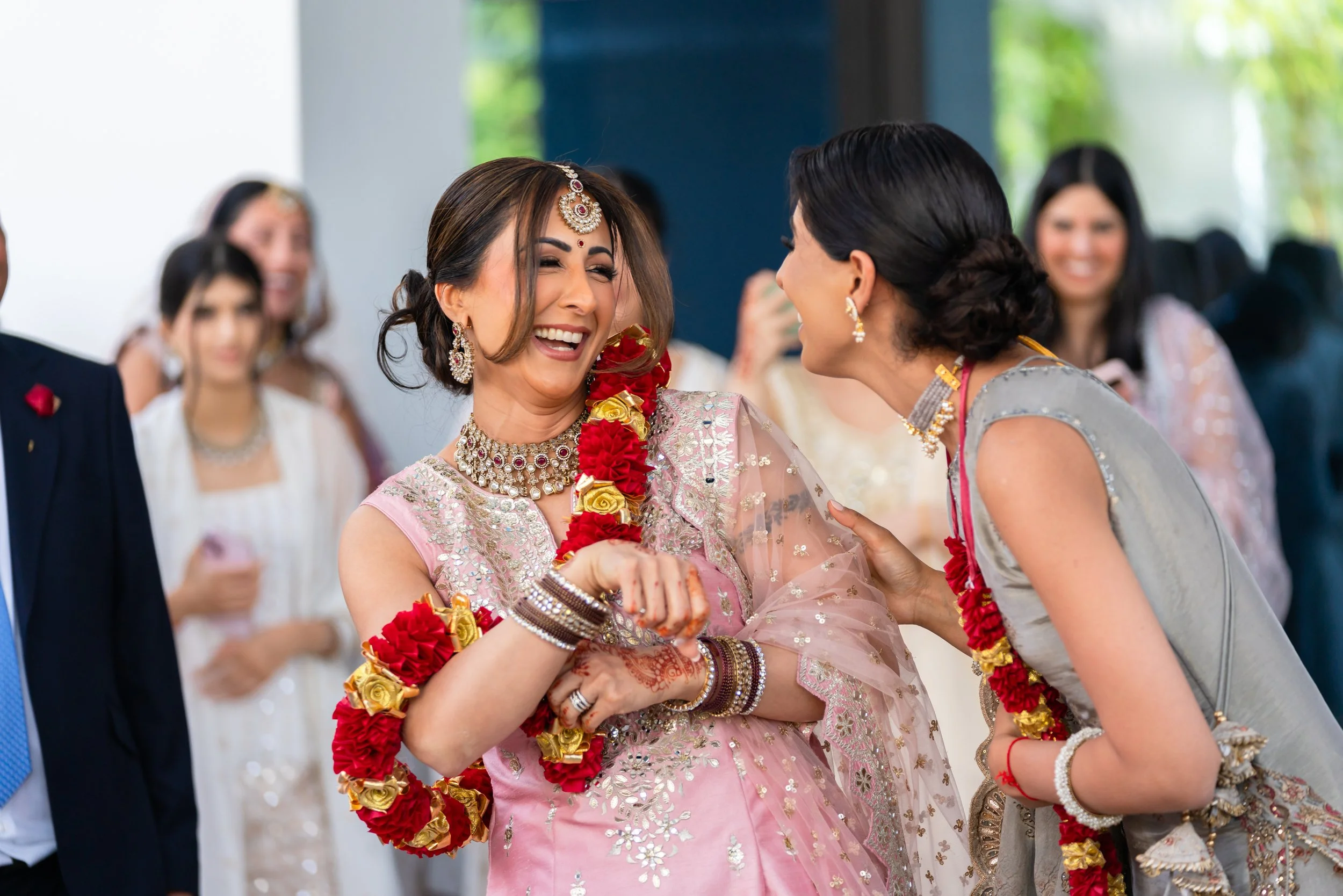 Two women in traditional Indian attire laughing and talking at a celebration, with other people in the background.