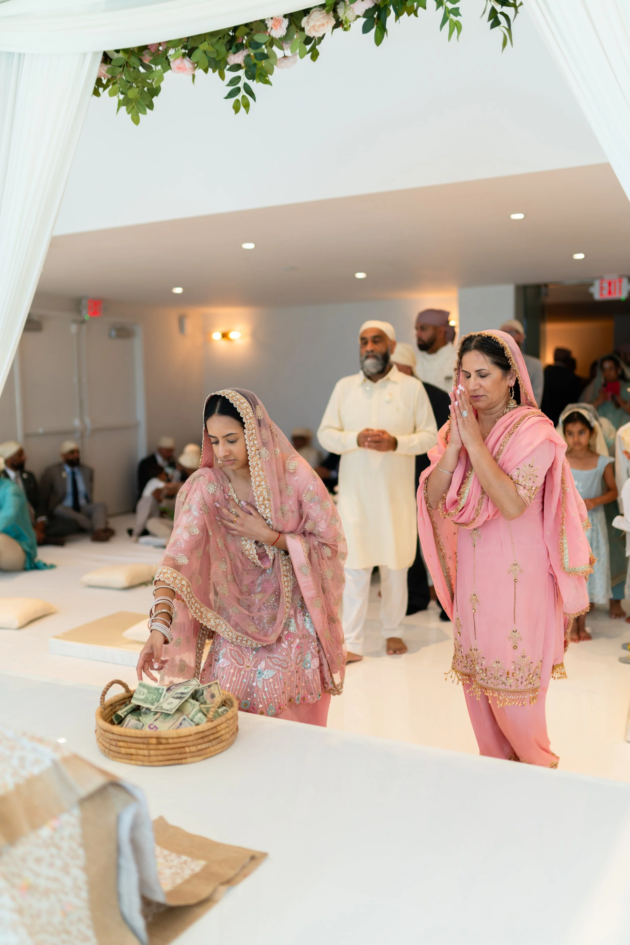 Women dressed in pink traditional Indian attire participating in a religious ceremony, with elders and children around in an indoor setting decorated with flowers.