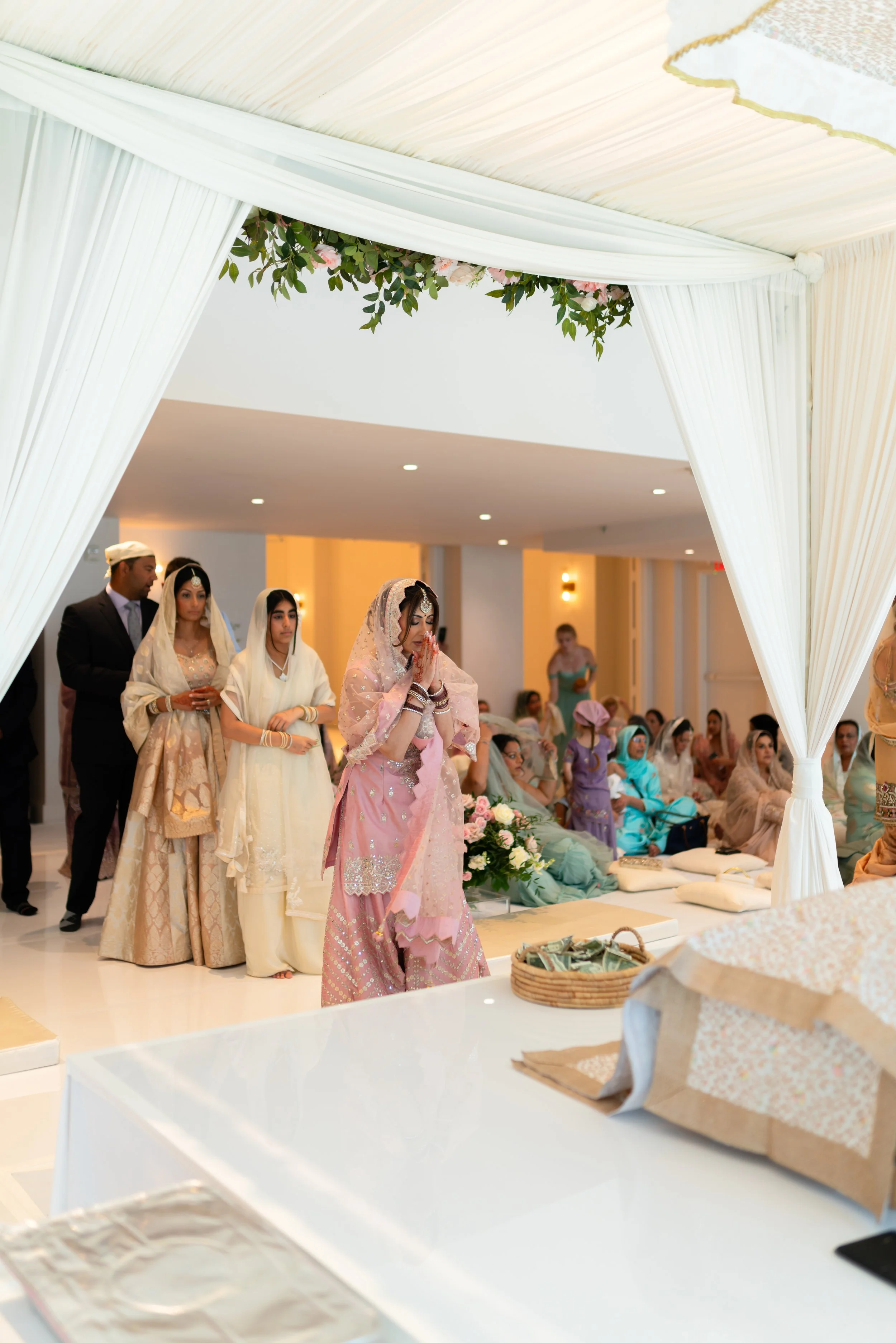 A traditional South Asian wedding ceremony with women dressed in colorful sarees and lehengas, seated on the floor under a decorated canopy with white drapes and floral arrangements, with guests watching.