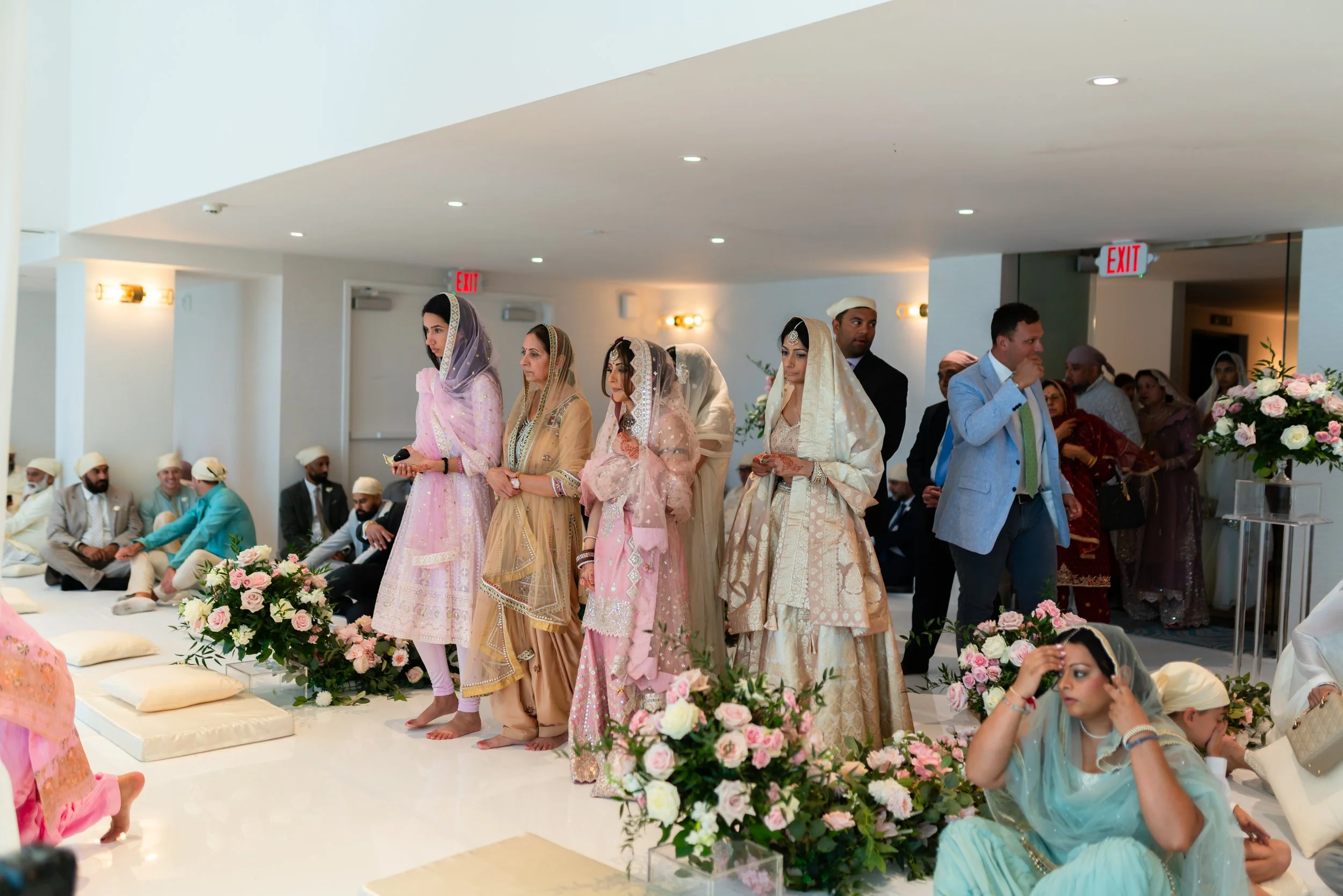 Indian wedding ceremony with women dressed in traditional bridal lehengas and men in traditional attire, sitting and standing around floral arrangements.