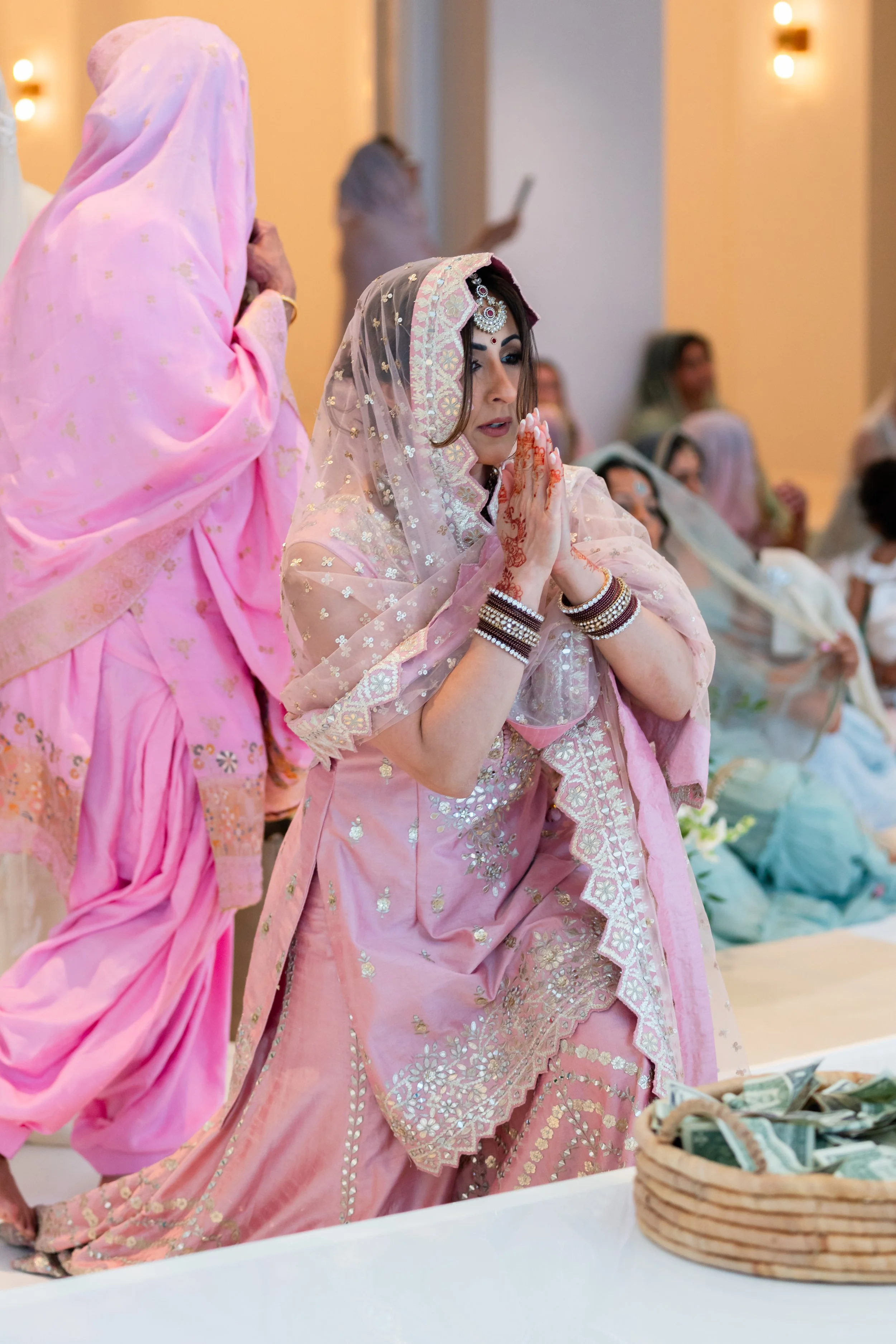 A woman dressed in traditional pink Indian attire with intricate embroidery, wearing jewelry and henna, praying at a wedding ceremony. She is kneeling with her hands folded and appears to be in a spiritual moment, surrounded by other women in colorfu