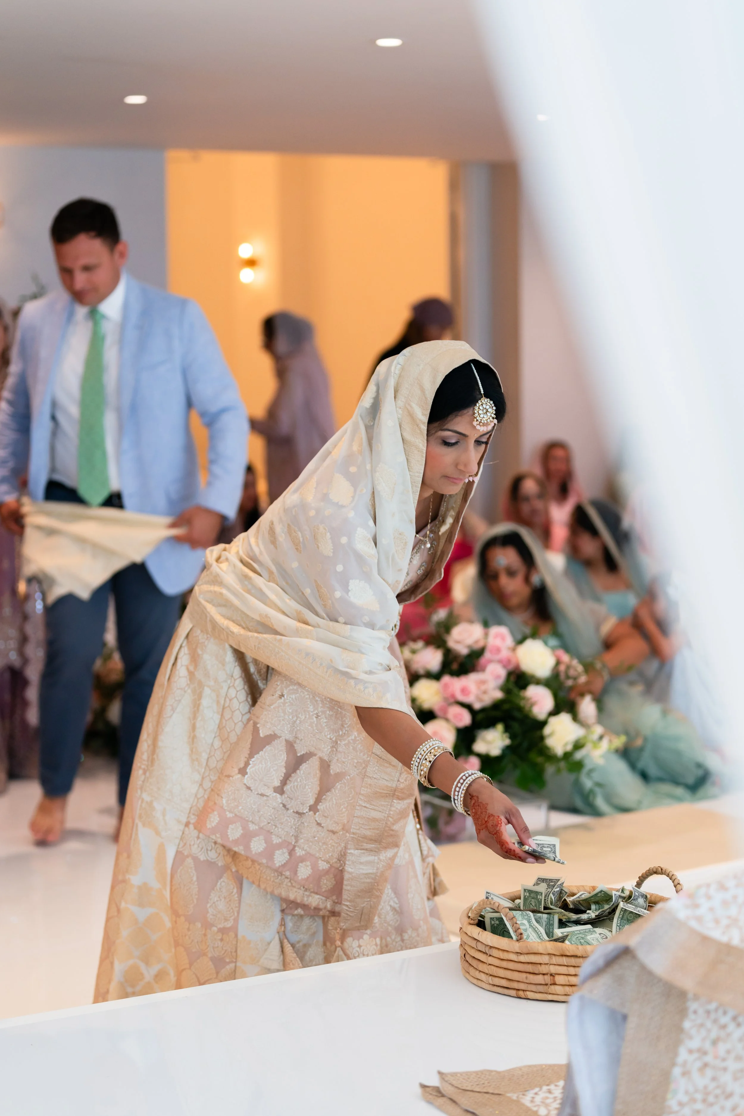 A woman dressed in traditional Indian attire placing money into a basket, with others sitting and standing around her in a festive setting.