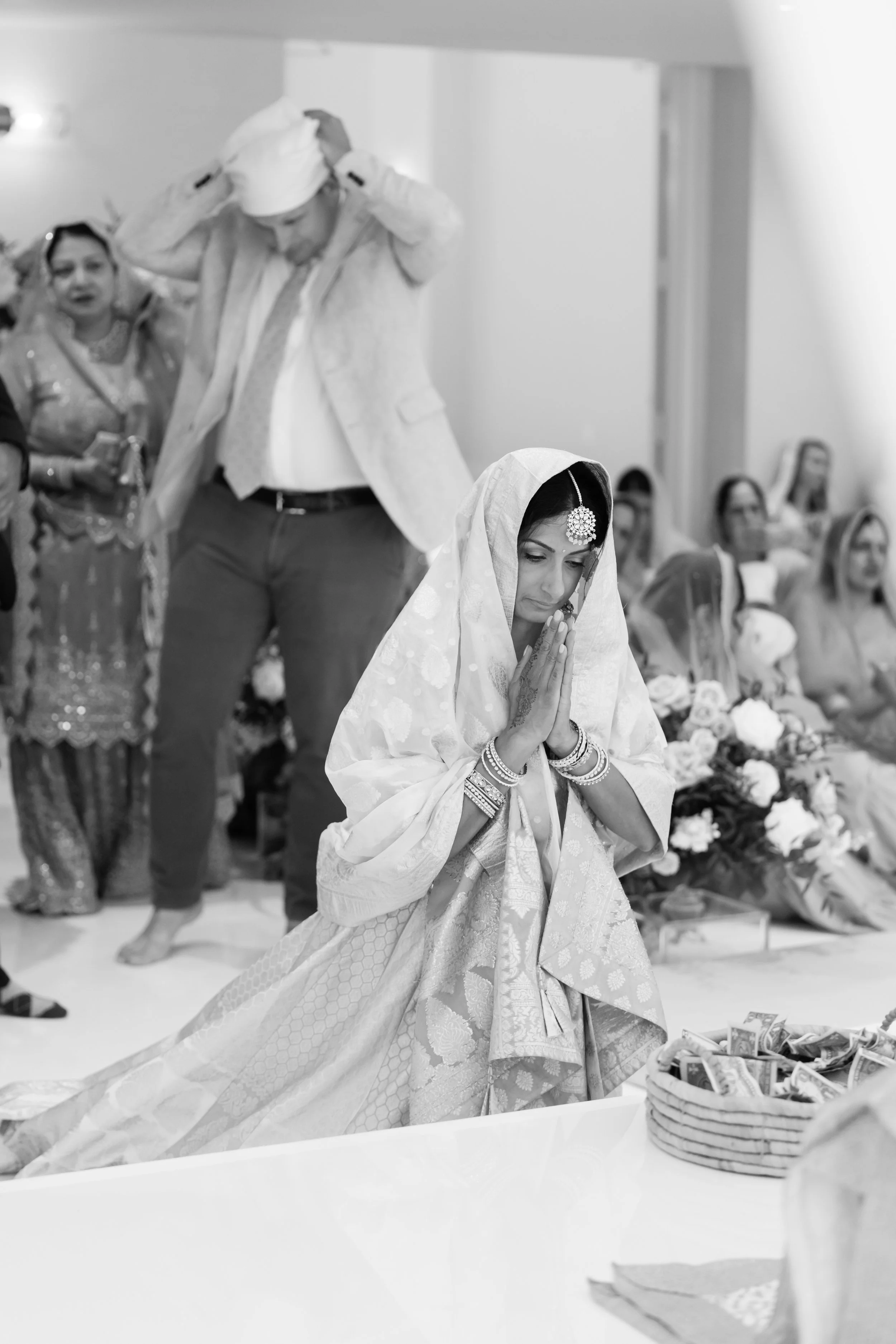 A woman dressed in traditional Indian attire with jewelry, praying with folded hands, during a cultural or religious event. People are seated in the background, and a basket of currency notes is visible on a table in front.