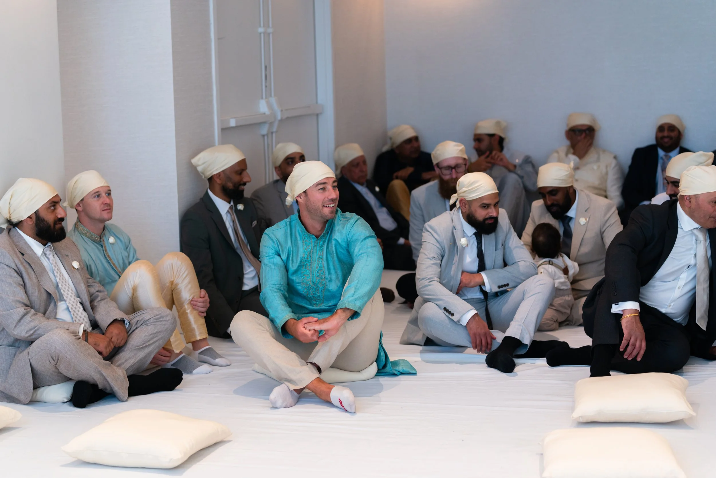 A group of men dressed in formal attire and traditional turbans, sitting on the floor during a religious or cultural ceremony.